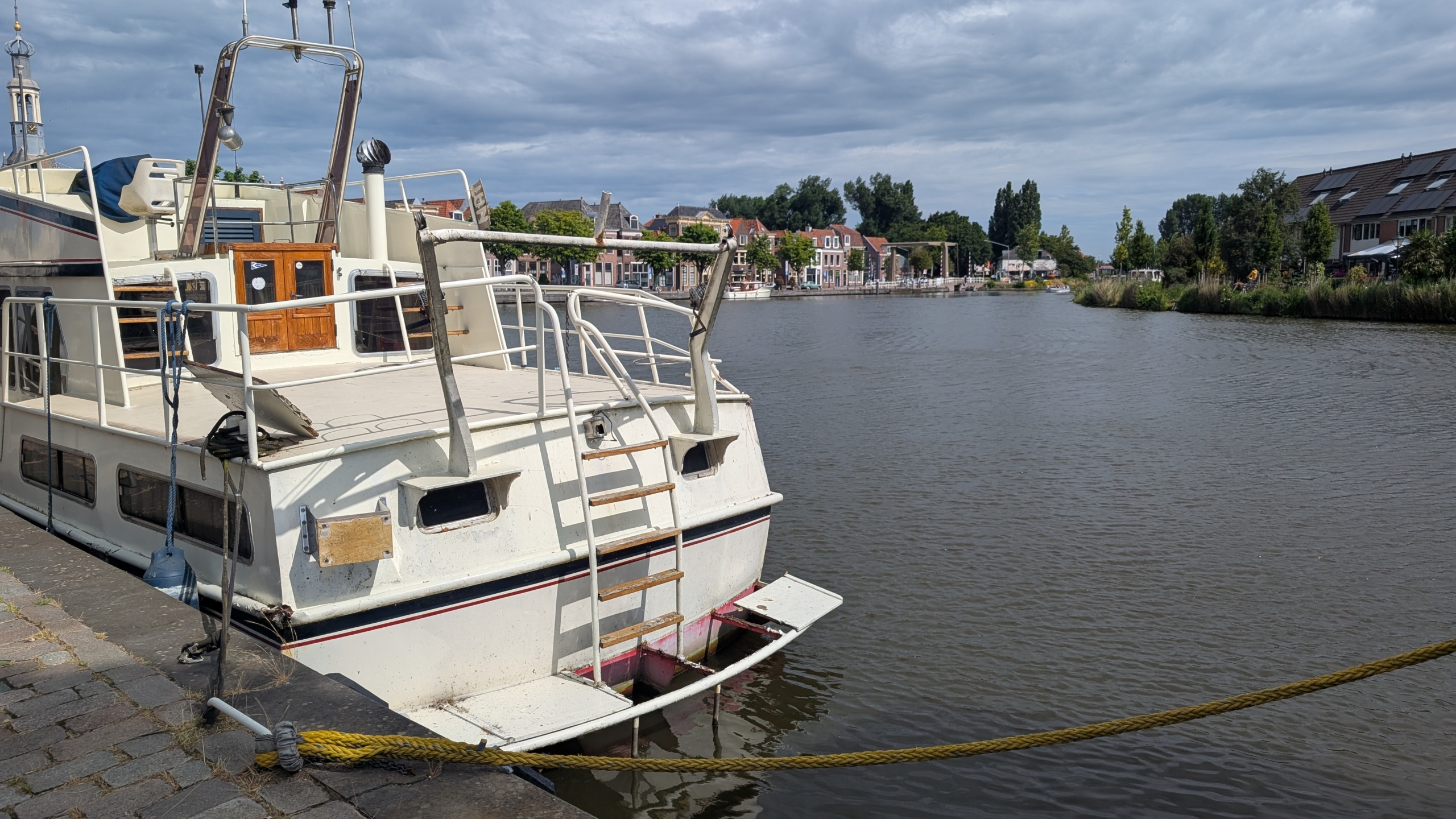 Witte boot aangemeerd langs een kanaal met huizen en bomen op de achtergrond onder een bewolkte hemel.