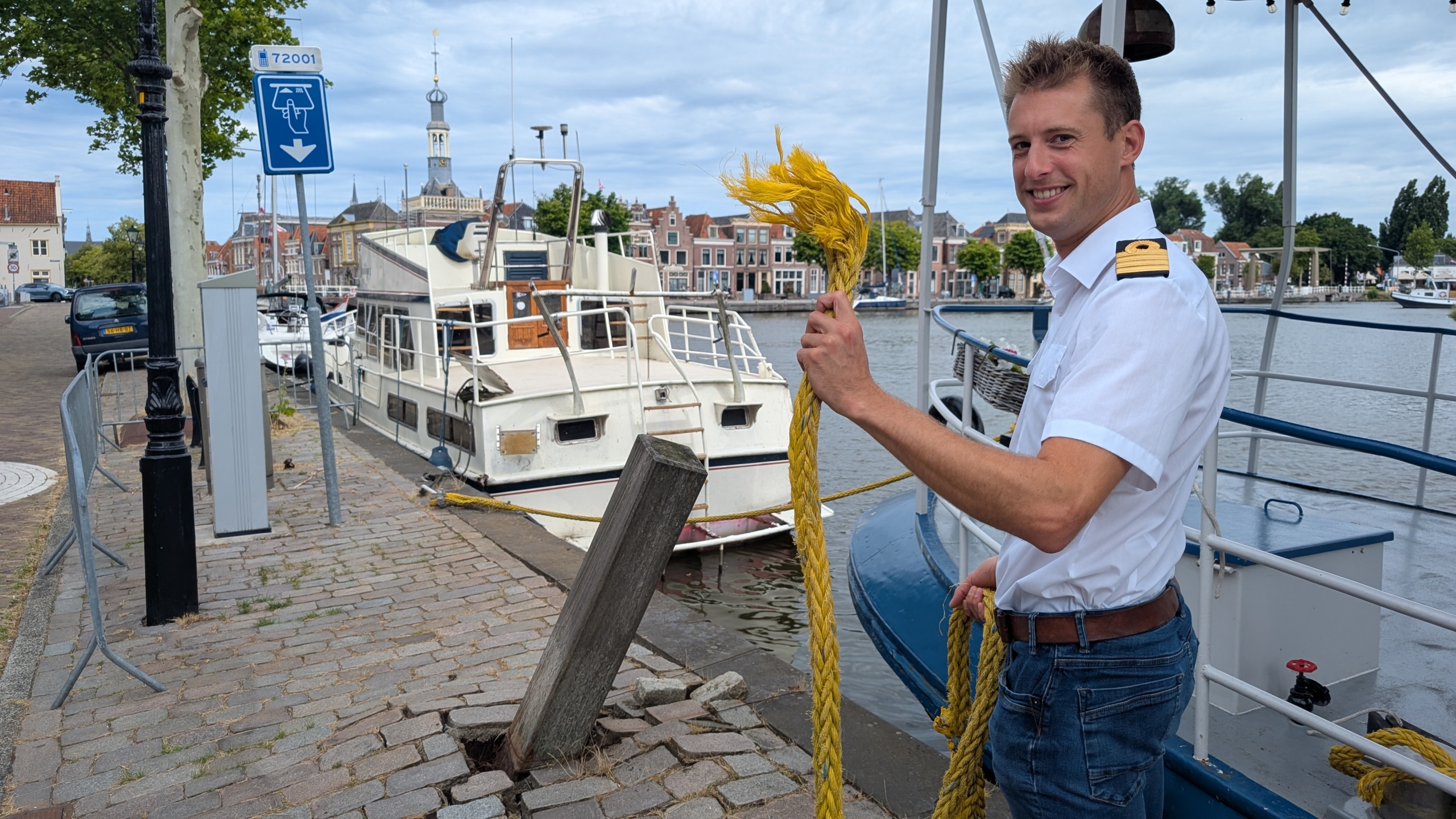 Een man in een wit uniform staat bij een kanaal met een gele touw, naast een aangemeerde boot.