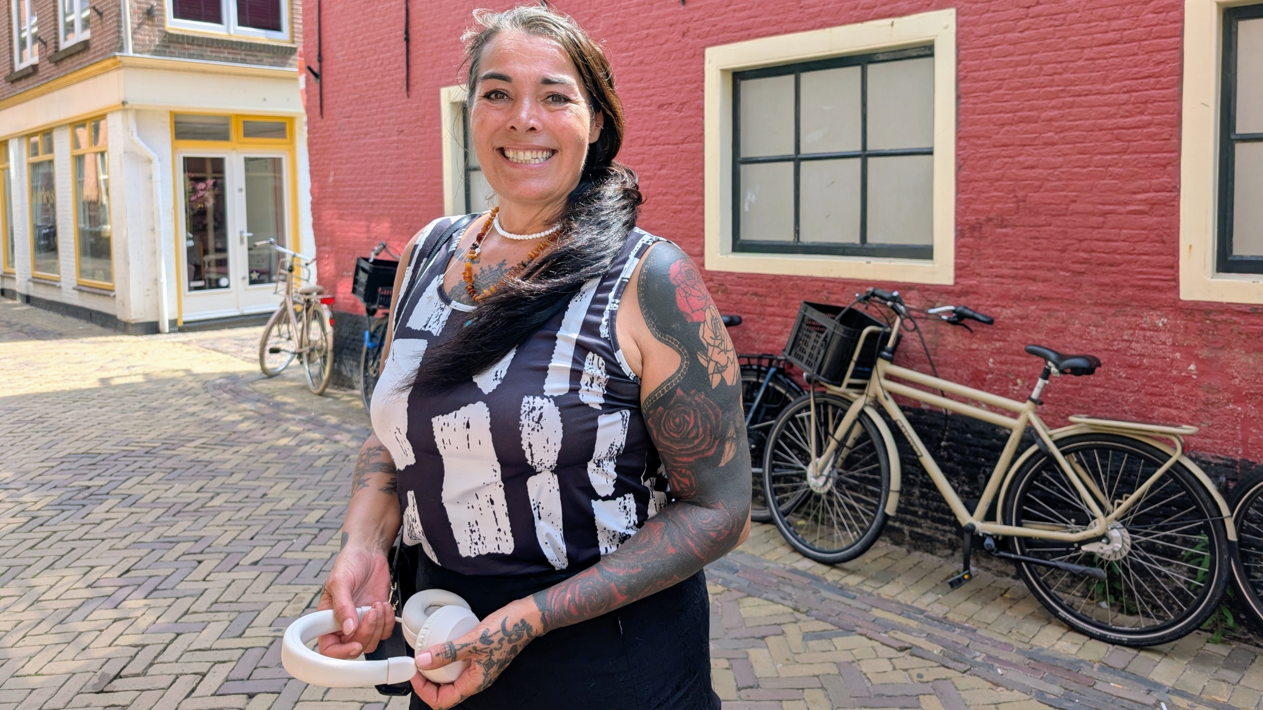 Vrouw met getatoeëerde arm glimlacht buiten naast fiets en rode bakstenen muur, met hoofdtelefoon in haar hand.