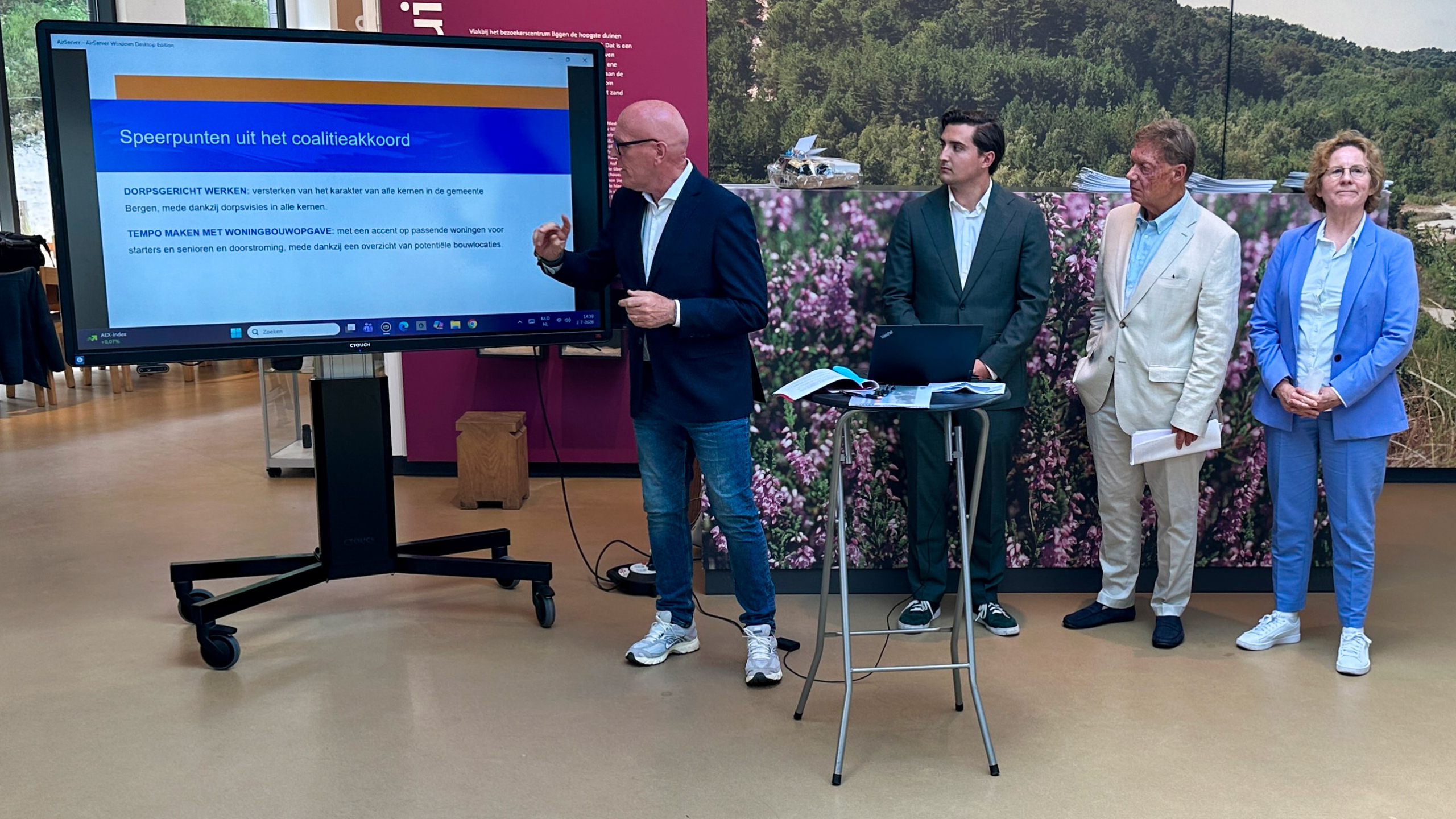 Man presenteert speerpunten uit het coalitieakkoord op een scherm, terwijl drie andere personen toekijken in een zaal.