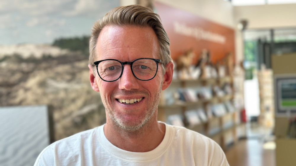Een man met een bril en kort blond haar glimlacht in een lichte ruimte met planken vol boeken.