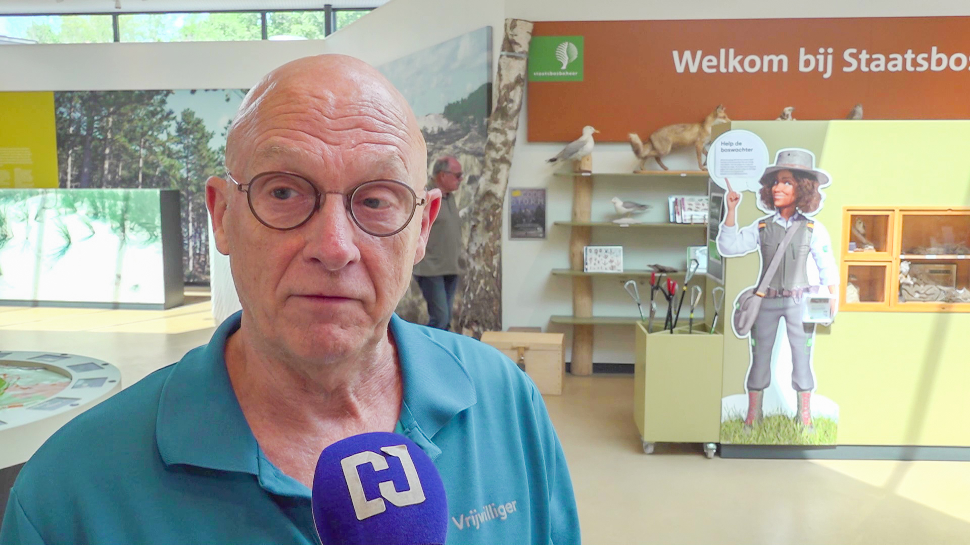 Een man met een bril wordt geïnterviewd in een bezoekerscentrum met op de achtergrond een bord "Welkom bij Staatsbosbeheer".