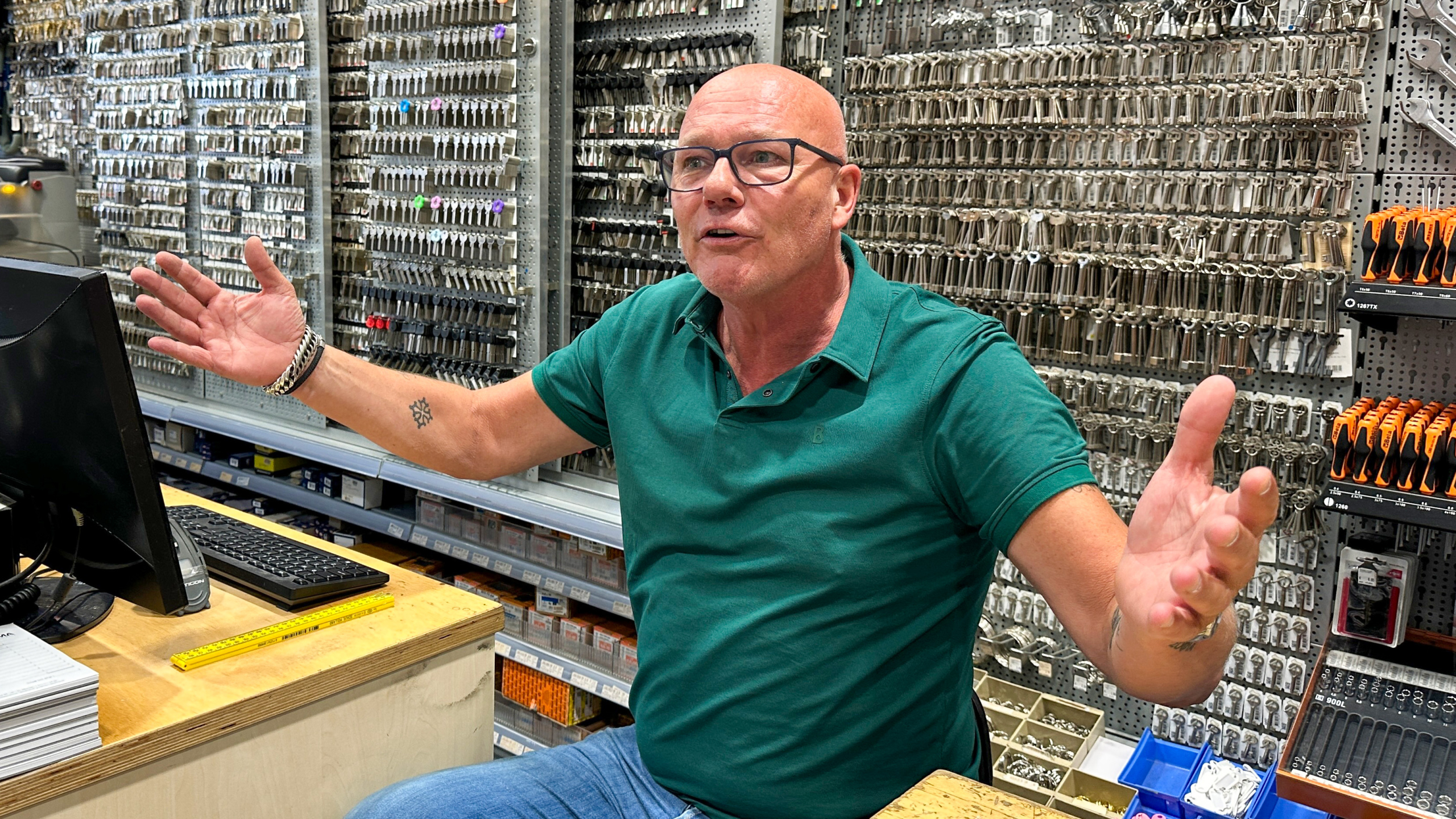 Man in een groen poloshirt, sprekend achter een balie in een winkel vol met sleutels aan de muur.