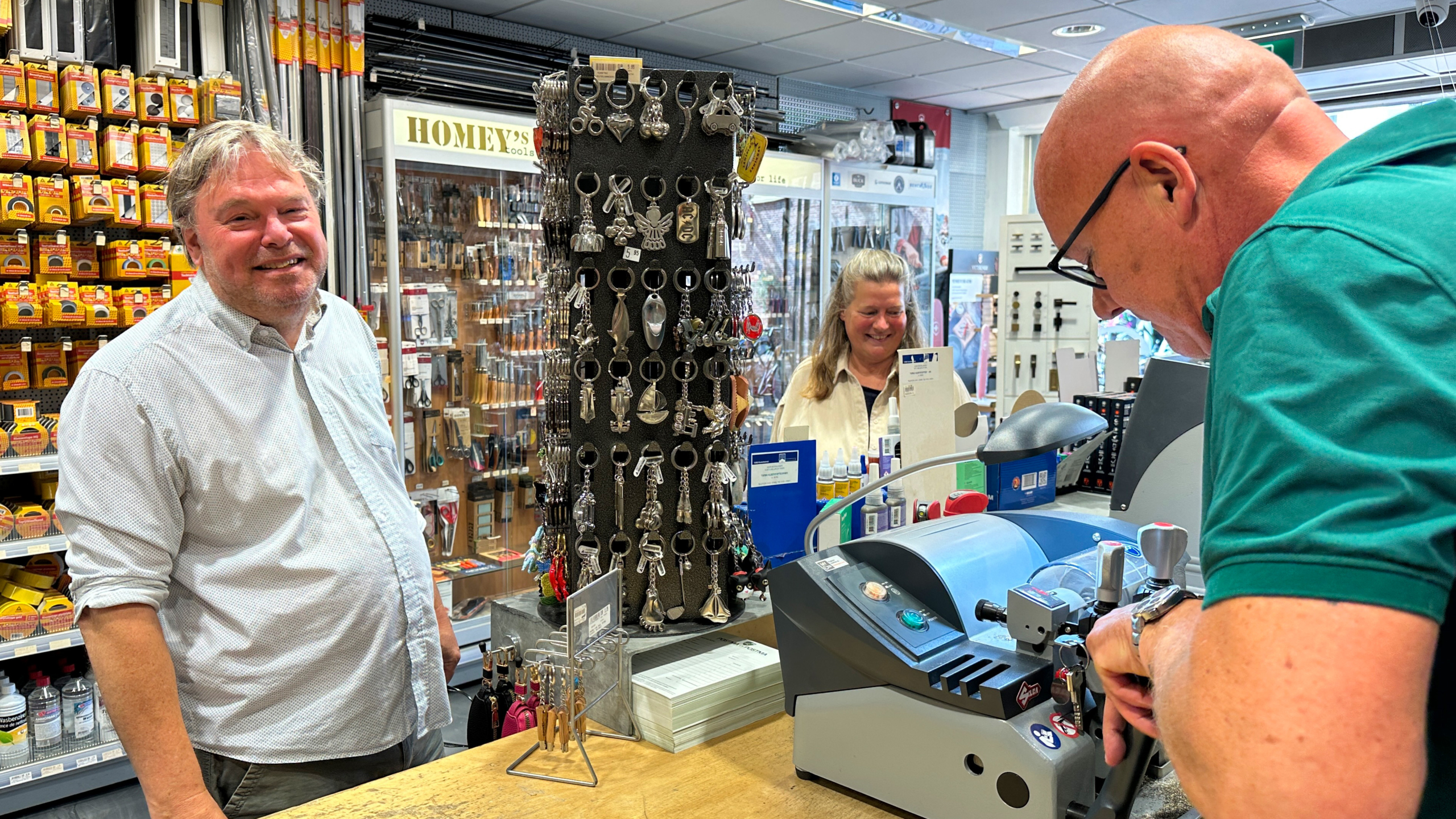 Mensen in een winkel bij een balie met een sleutelslijpmachine, een sleutelhangerdisplay en diverse gereedschappen op de achtergrond.