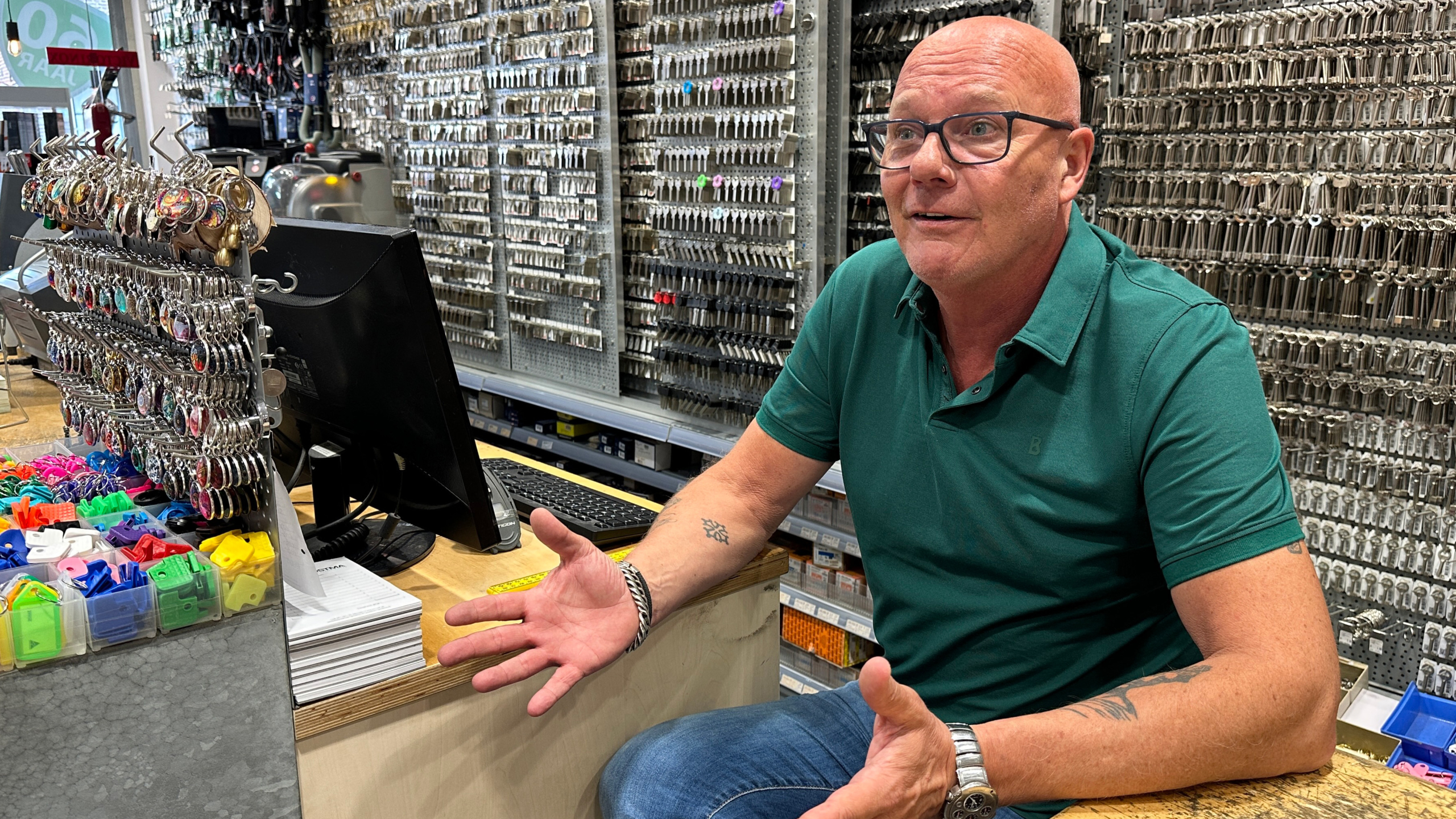 Man bij een balie in een winkel vol sleutels, pratend en gebarend, omringd door diverse sleutelhangers en computer.