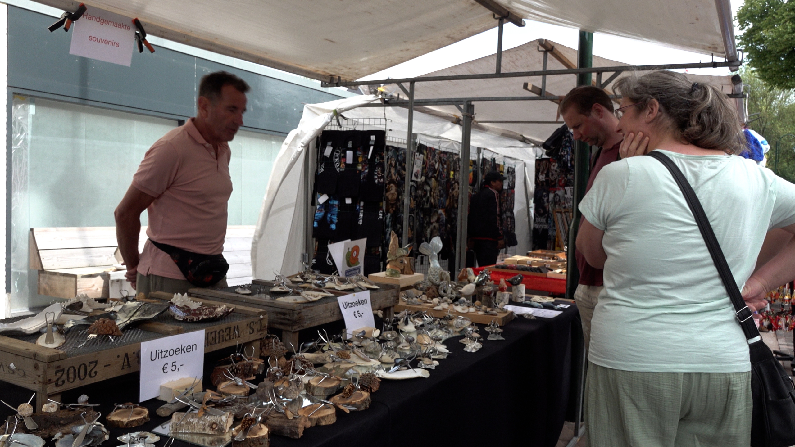 Een man verkoopt handgemaakte souvenirs op een markt, terwijl twee mensen de uitgestalde producten bekijken.