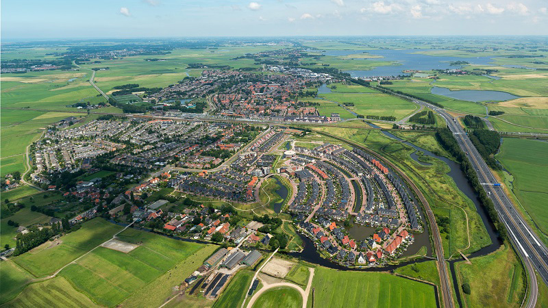 Luchtfoto van een uitgestrekt groen landschap met een woonwijk. Slingerweg en waterwegen zijn zichtbaar tussen de huizen.
