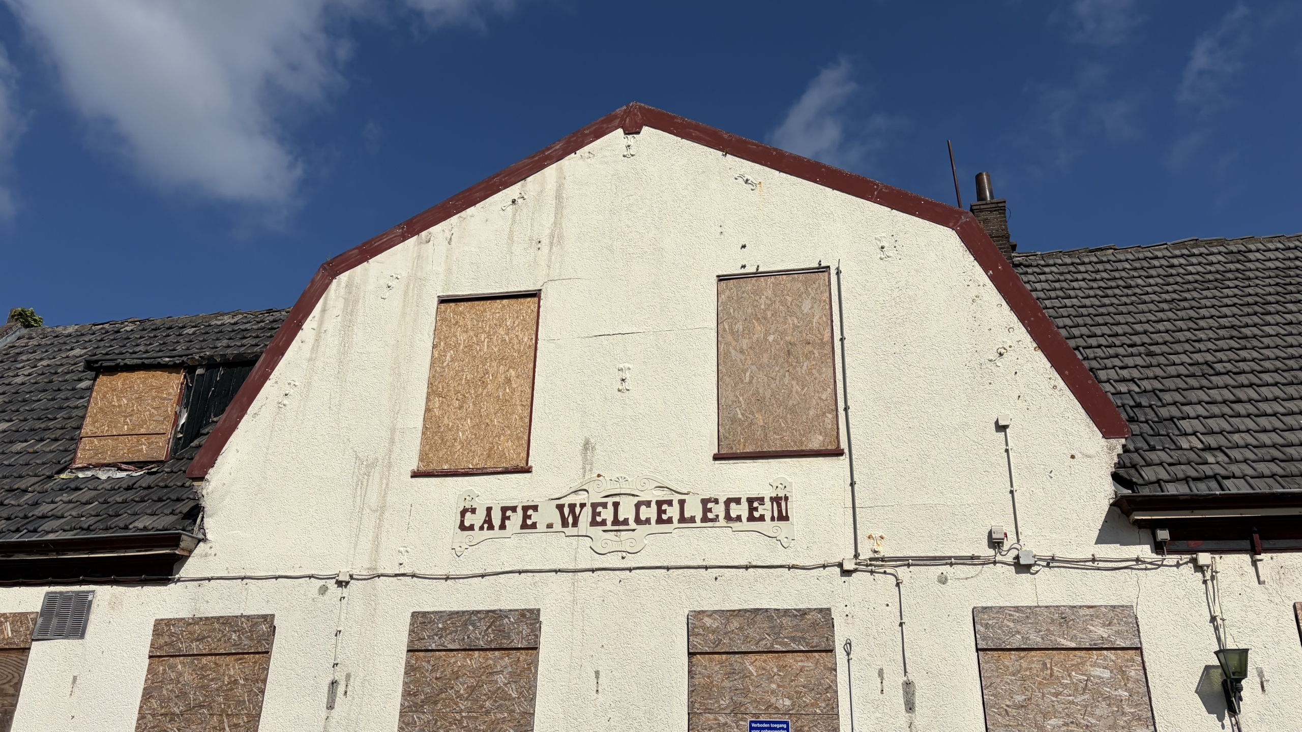 Een verlaten gebouw met dichtgetimmerde ramen, met de tekst "Cafe Welcelecen" op de gevel onder een heldere blauwe hemel.