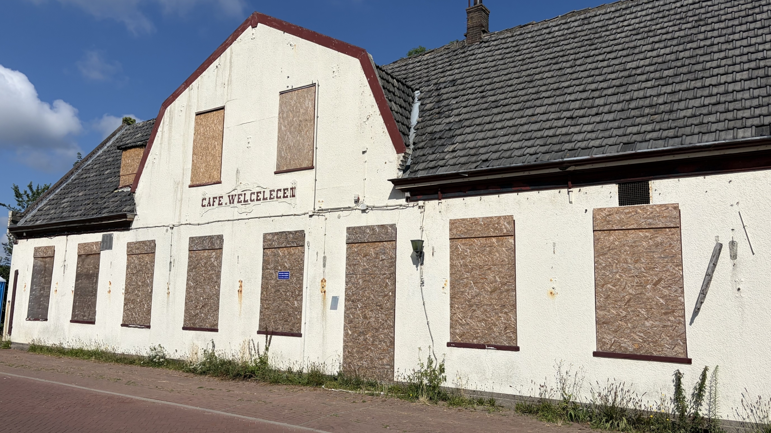 Oud gebouw van Café Welgelegen met dichtgetimmerde ramen en roestvlekken, naast een blauwe mobiele toiletunit.