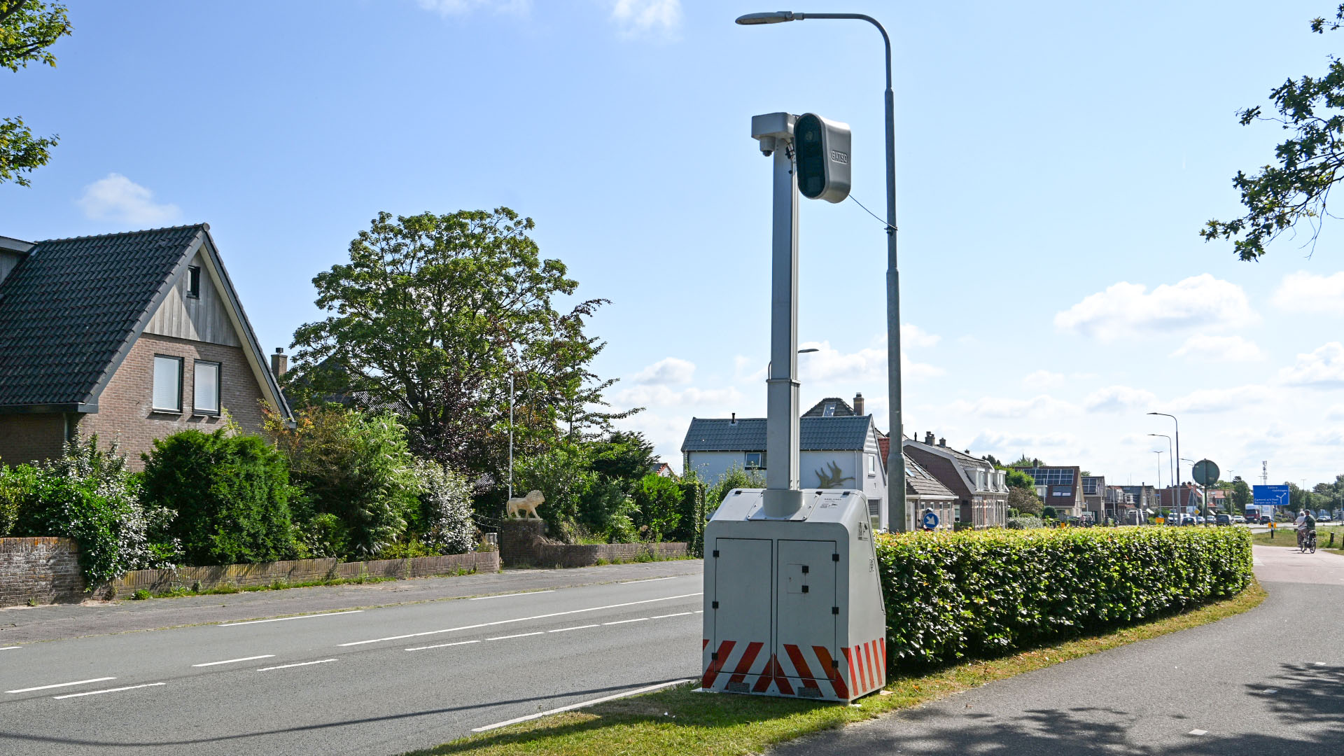 Mobiele flitspaal langs een weg in een woonwijk, met huizen en groen op de achtergrond