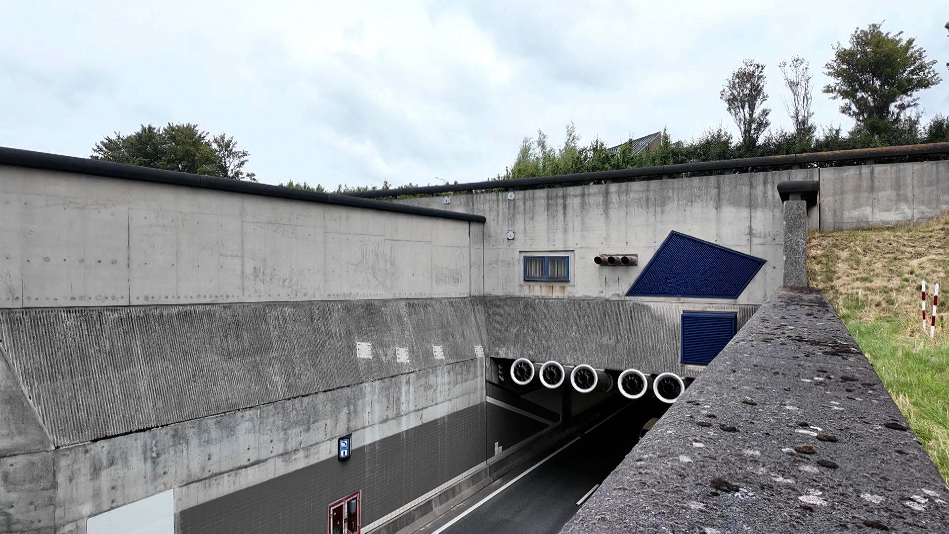 Ingang van een verkeerstunnel met betonnen wanden, blauwe ventilatieroosters en afwateringsbuizen onder een bewolkte lucht