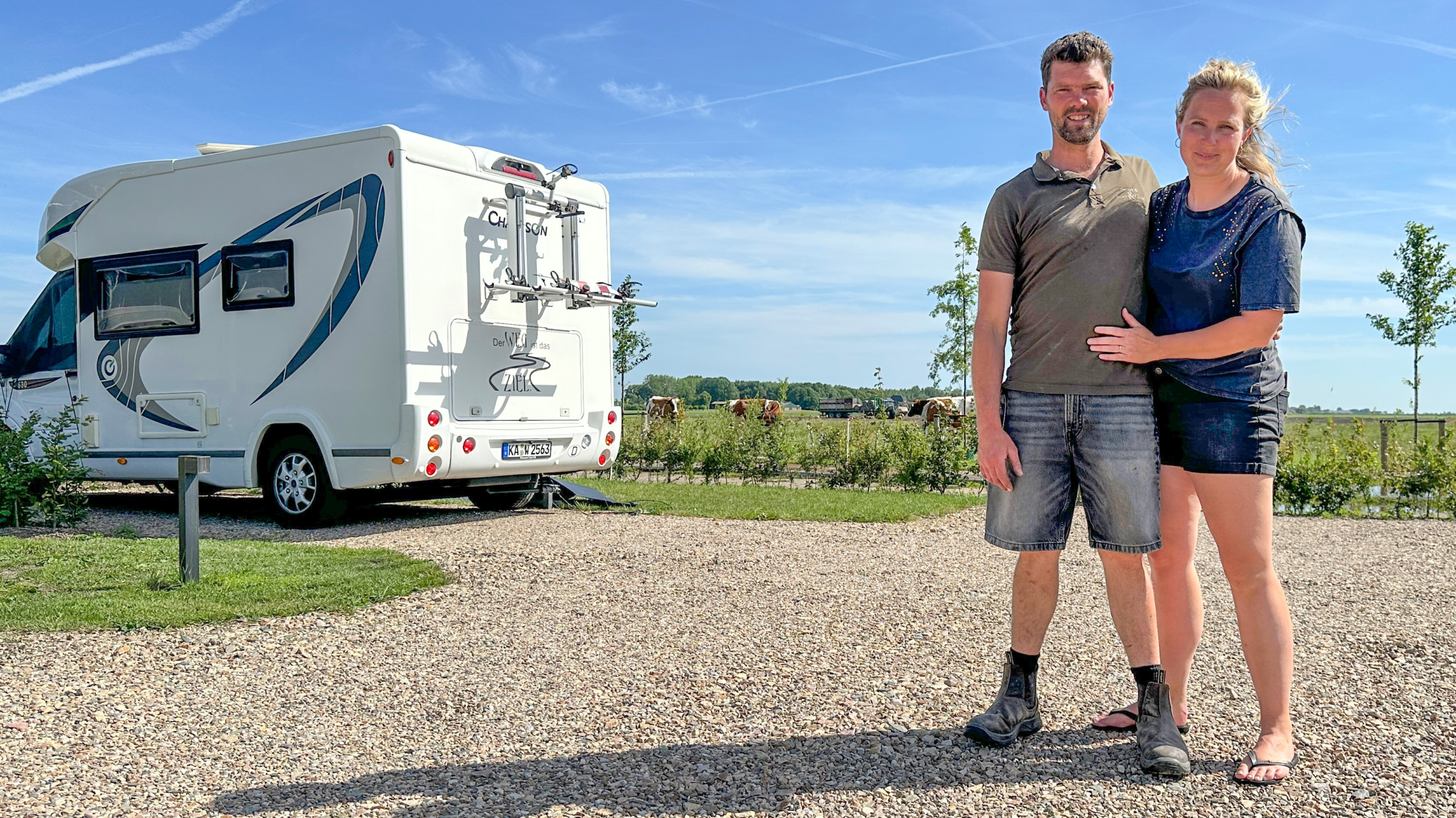 Stel poseert naast een witte camper op een camping, met weiland en koeien op de achtergrond onder blauwe lucht
