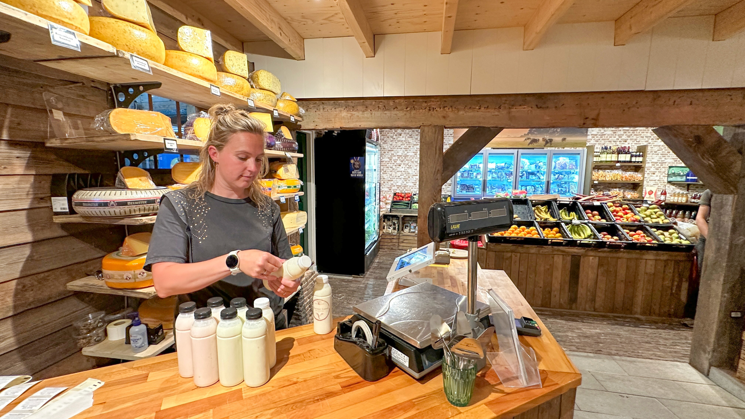 Medewerkster vult flessen zuivel bij de kassa in een boerderijwinkel met kazen op planken en groente- en fruitbakken