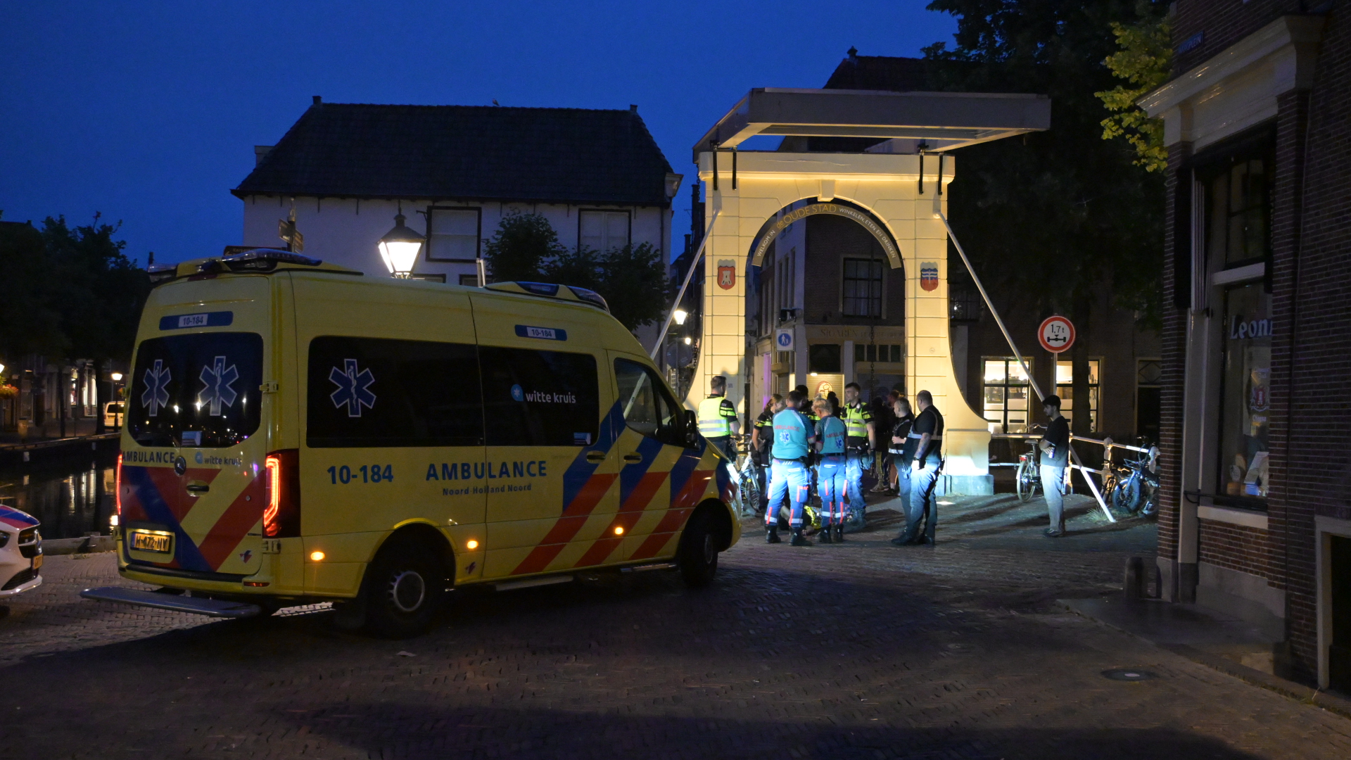Gele ambulance en hulpverleners bij een verlichte ophaalbrug in de avond, met omstanders op straat