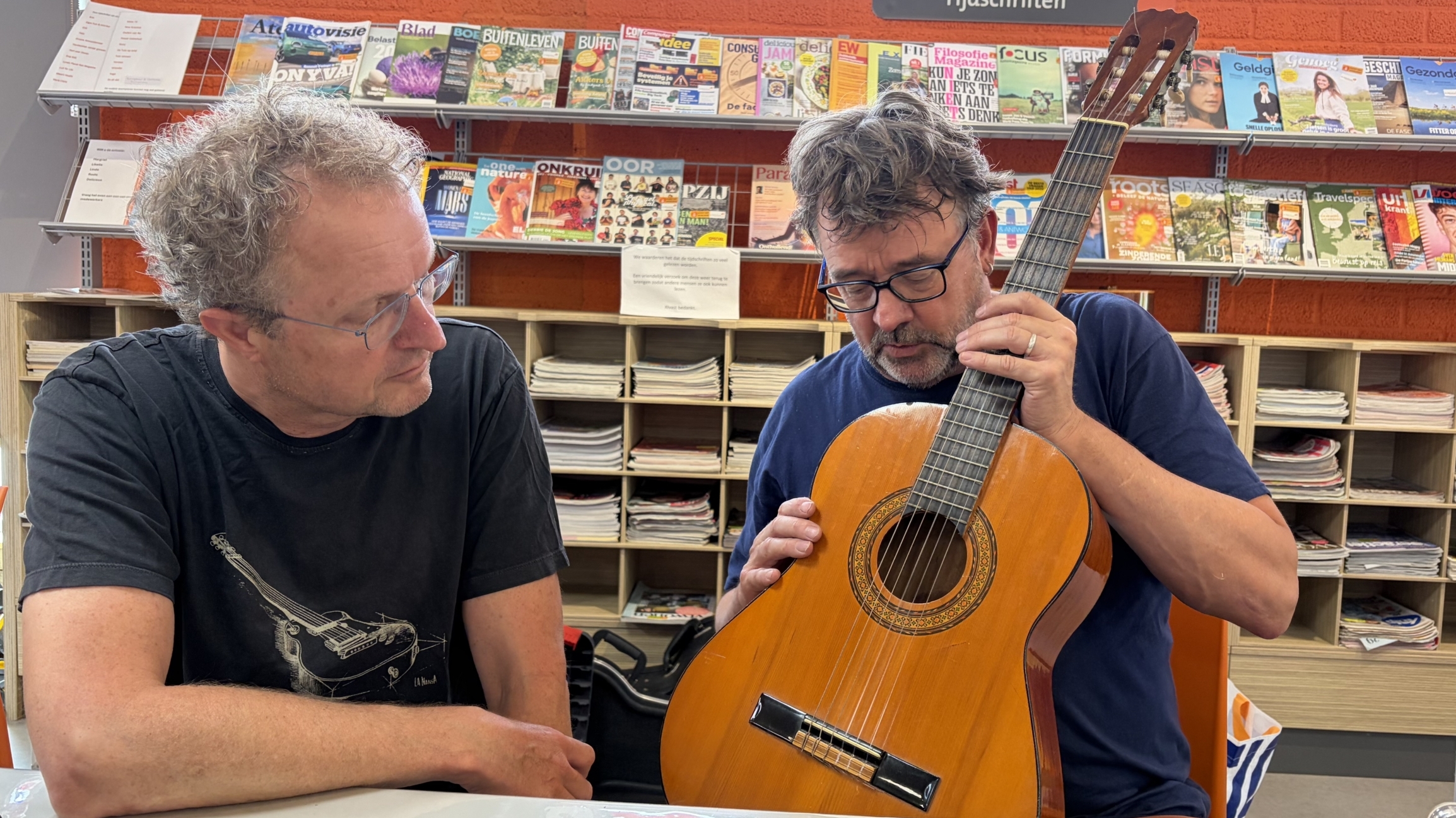 Twee mannen aan een tafel, één houdt een klassieke gitaar vast terwijl de ander toekijkt, met tijdschriftenrekken op de achtergrond