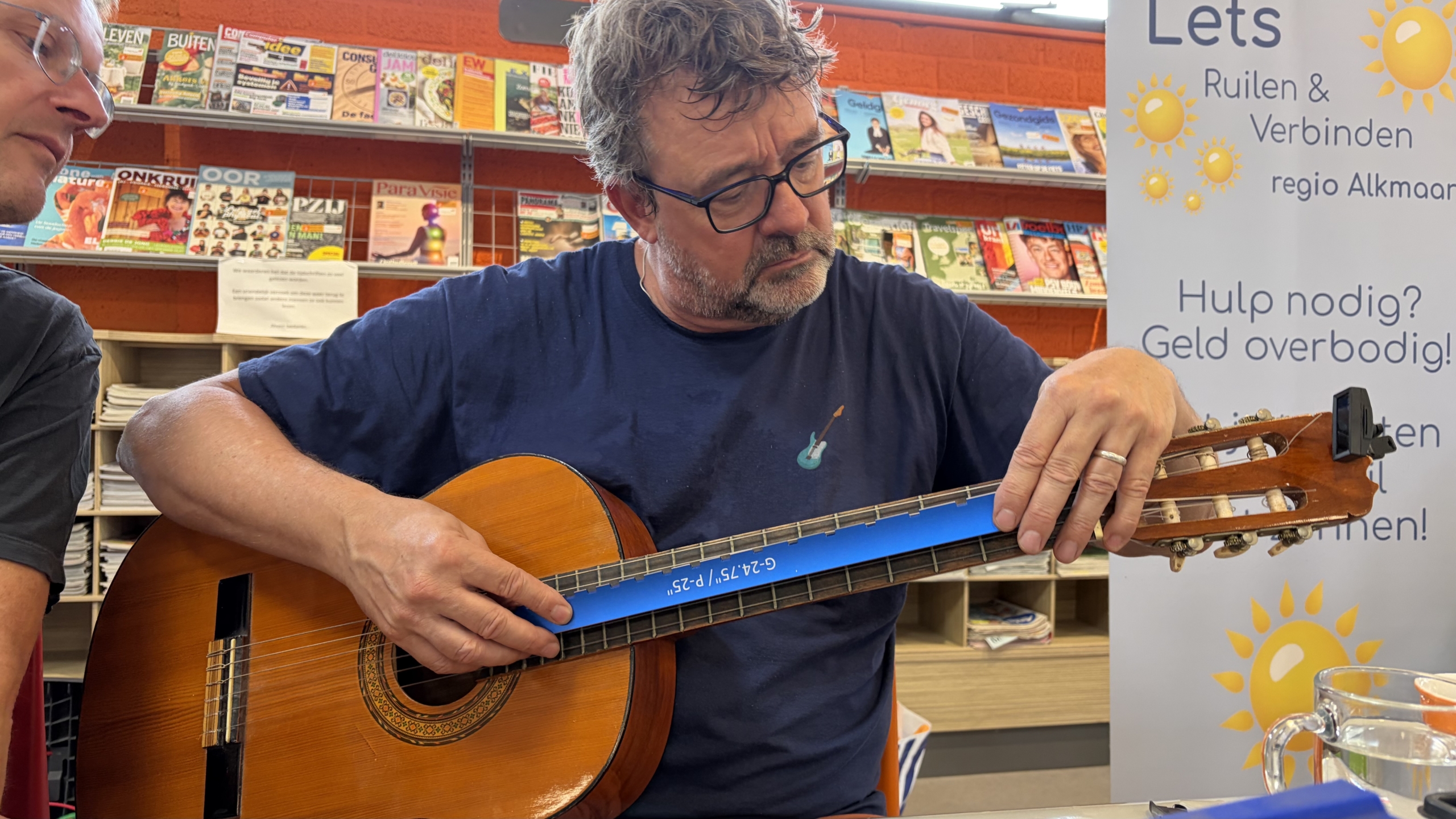 Man stemt een klassieke gitaar met een digitale tuner, met tijdschriftenrek en Lets Ruilen & Verbinden-banner op de achtergrond