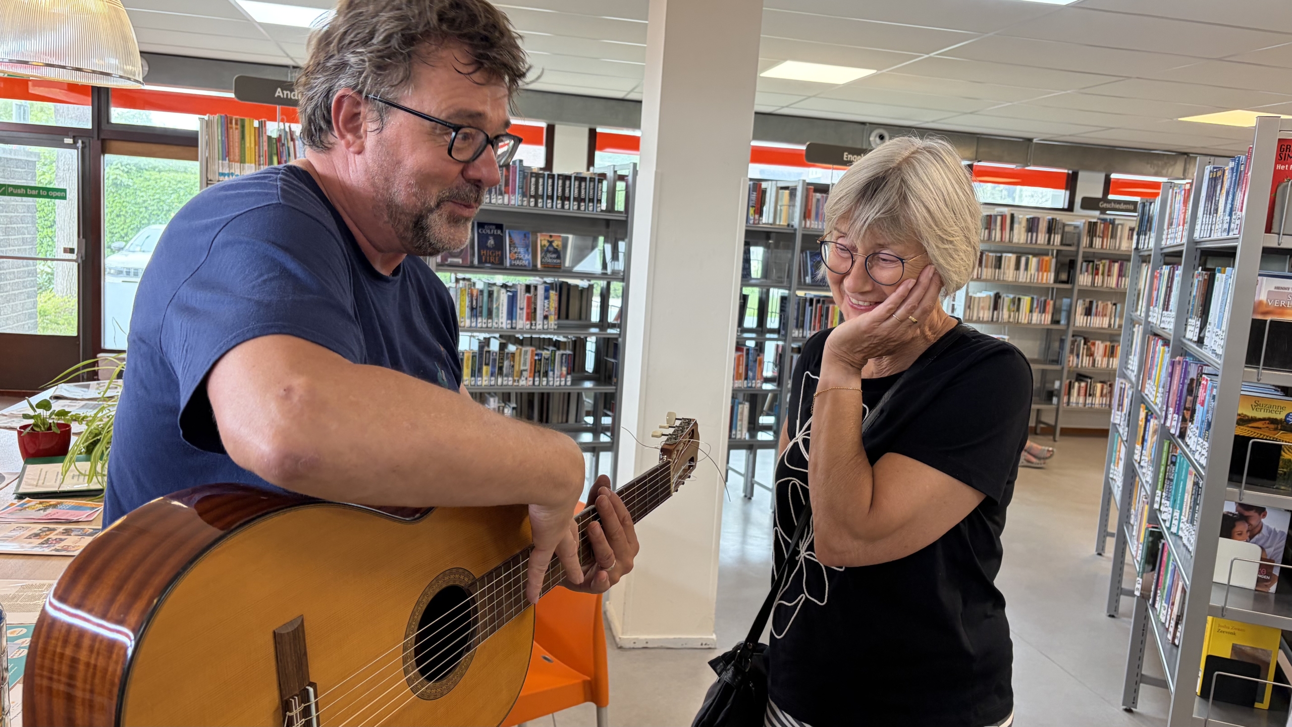 Man met bril houdt een akoestische gitaar vast terwijl een vrouw glimlachend luistert in een bibliotheek tussen boekenkasten