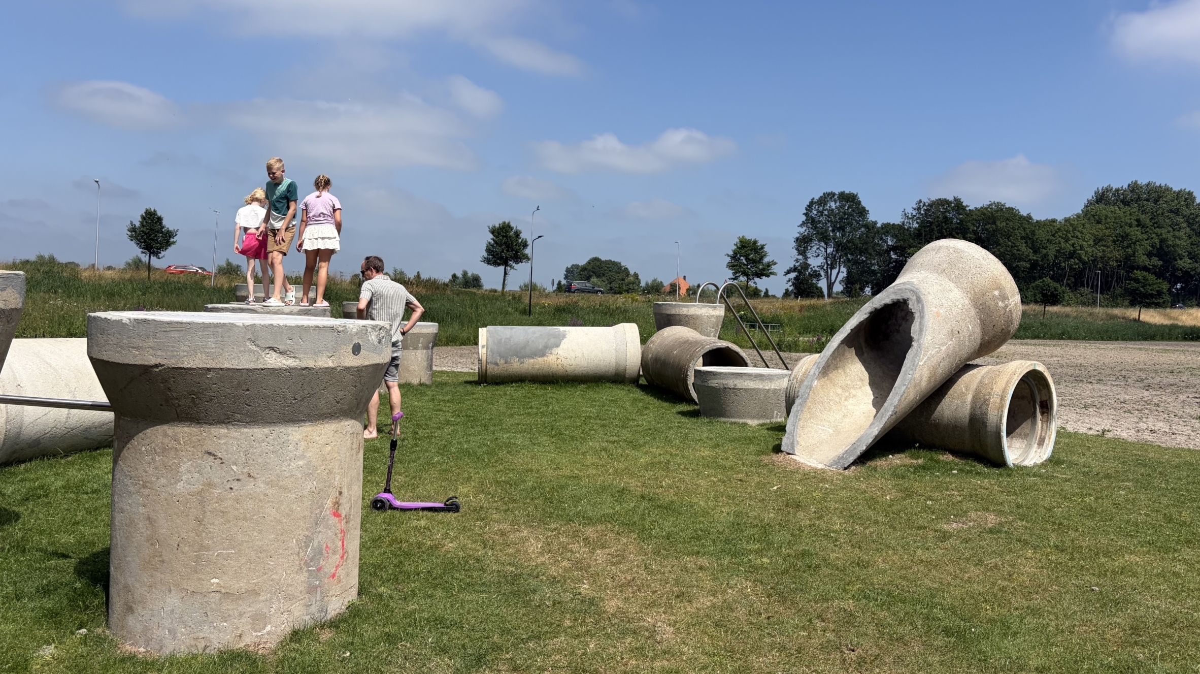 Kinderen spelen op grote betonnen buizen en ringen op een grasveld onder een blauwe lucht