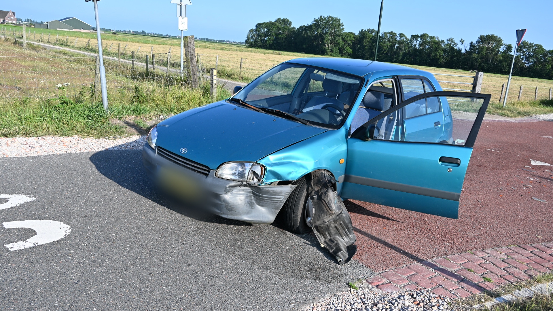 Blauwe auto met zware schade aan de linker voorkant staat op een kruispunt, met het rechterportier open