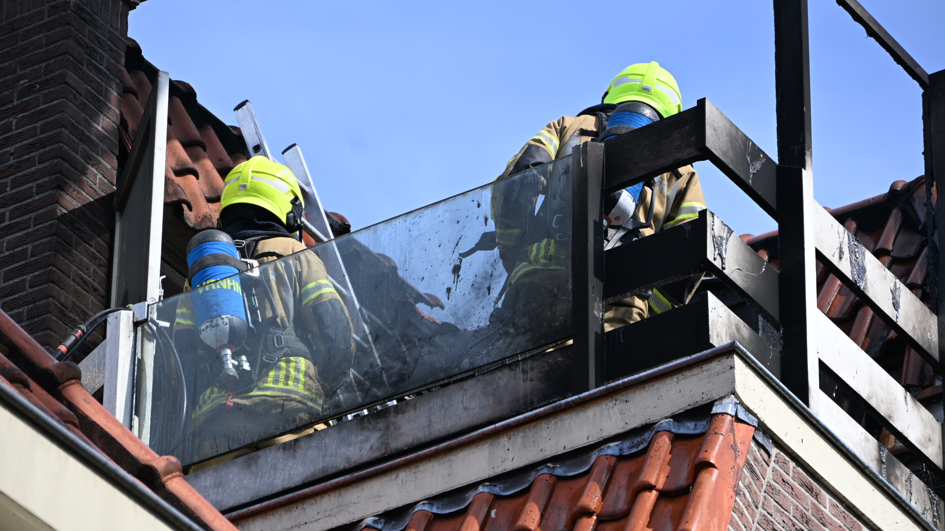 Twee brandweerlieden met gele helmen op een dakbalkon met ladder, bij een beschadigde schoorsteen en dakpannen