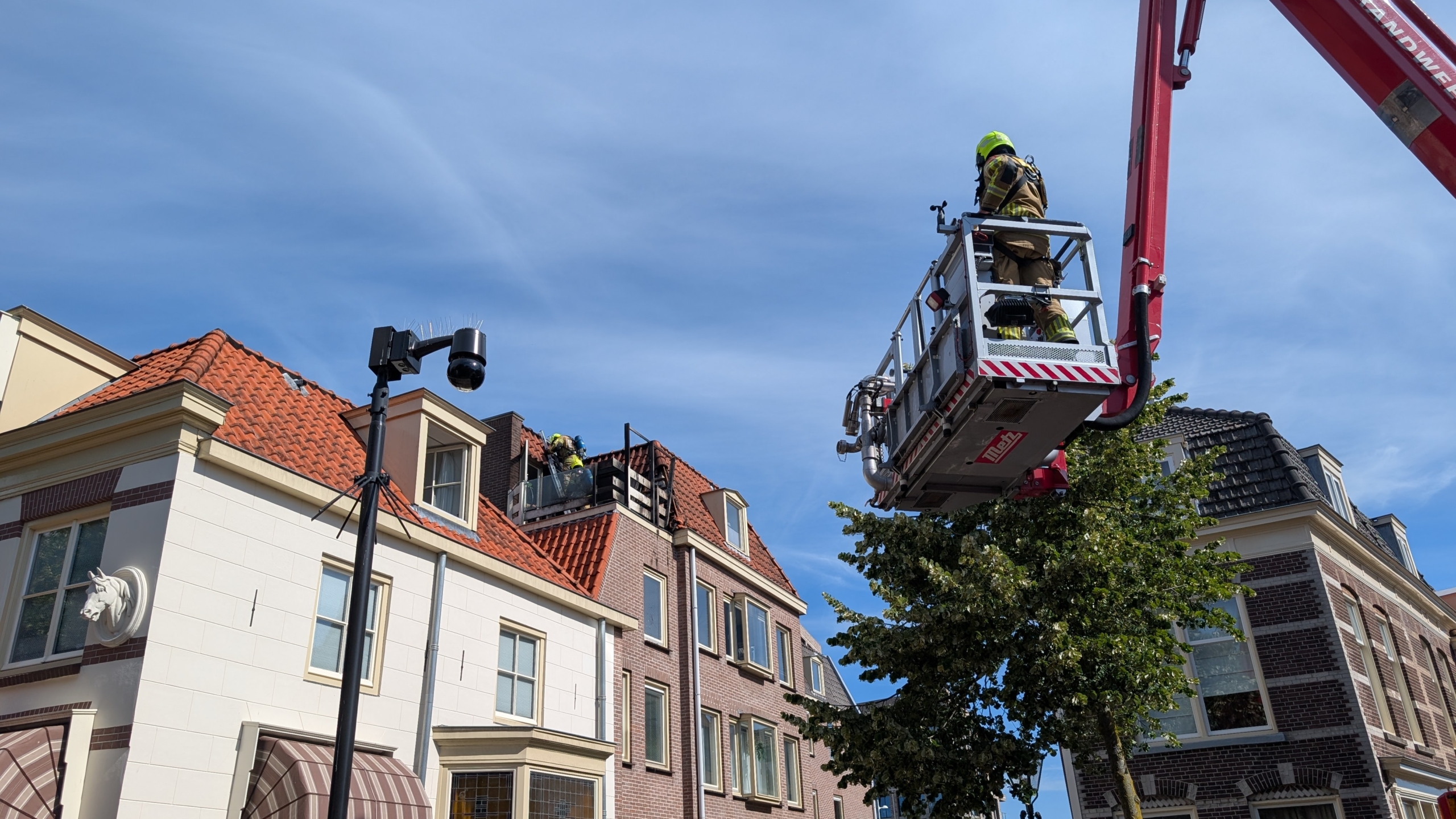 Brandweerman in een hoogwerker bij een dakkapel op een rood pannendak, met rook uit het dakraam