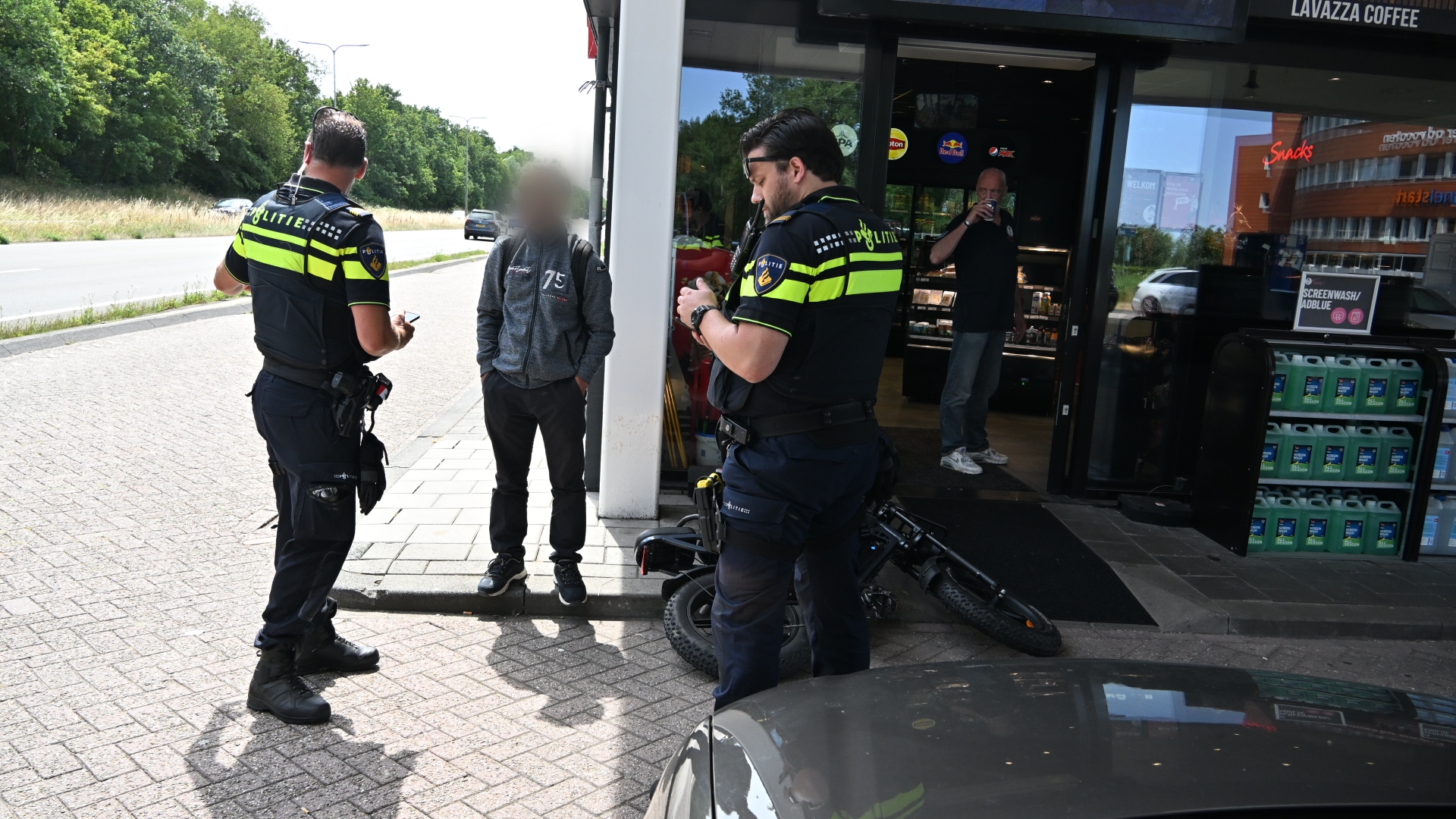 Twee politieagenten spreken een man aan bij een tankstationwinkel; een fiets ligt op de grond bij de ingang