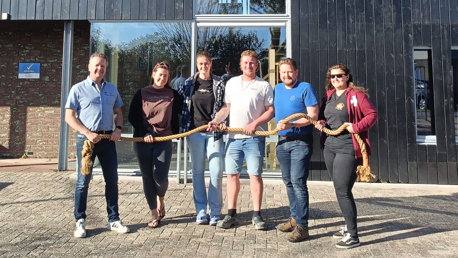 Zes mensen staan voor een gebouw en houden samen een dik touw vast, alsof ze aan touwtrekken doen