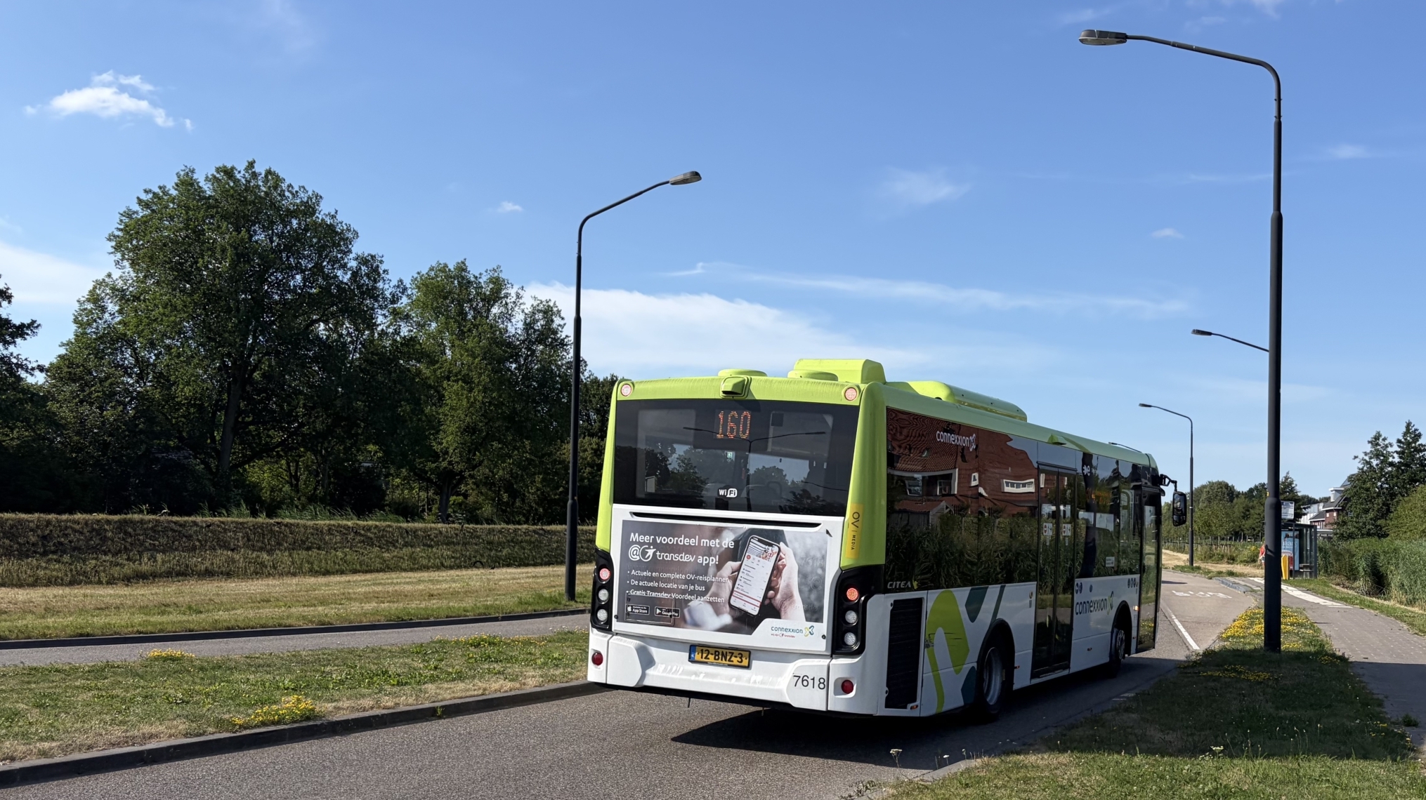 Groen-witte lijnbus rijdt over een rustige weg langs gras en bomen, met halte en straatlantaarns onder blauwe lucht