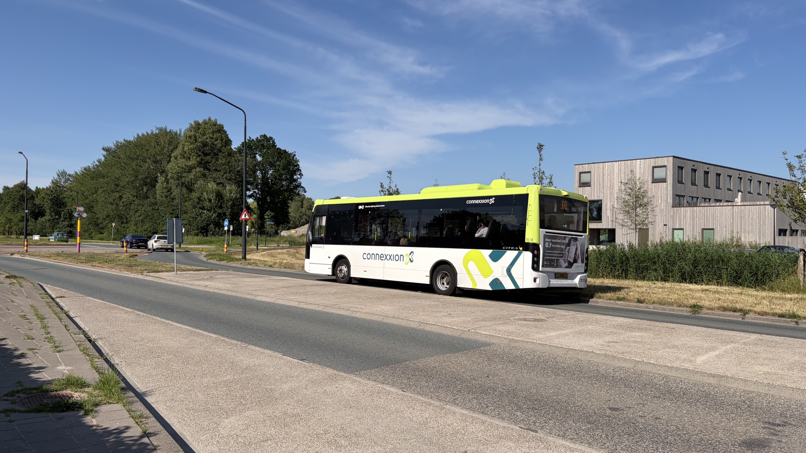 Connexxion-bus rijdt op een weg langs bomen en een modern gebouw, onder een heldere blauwe lucht