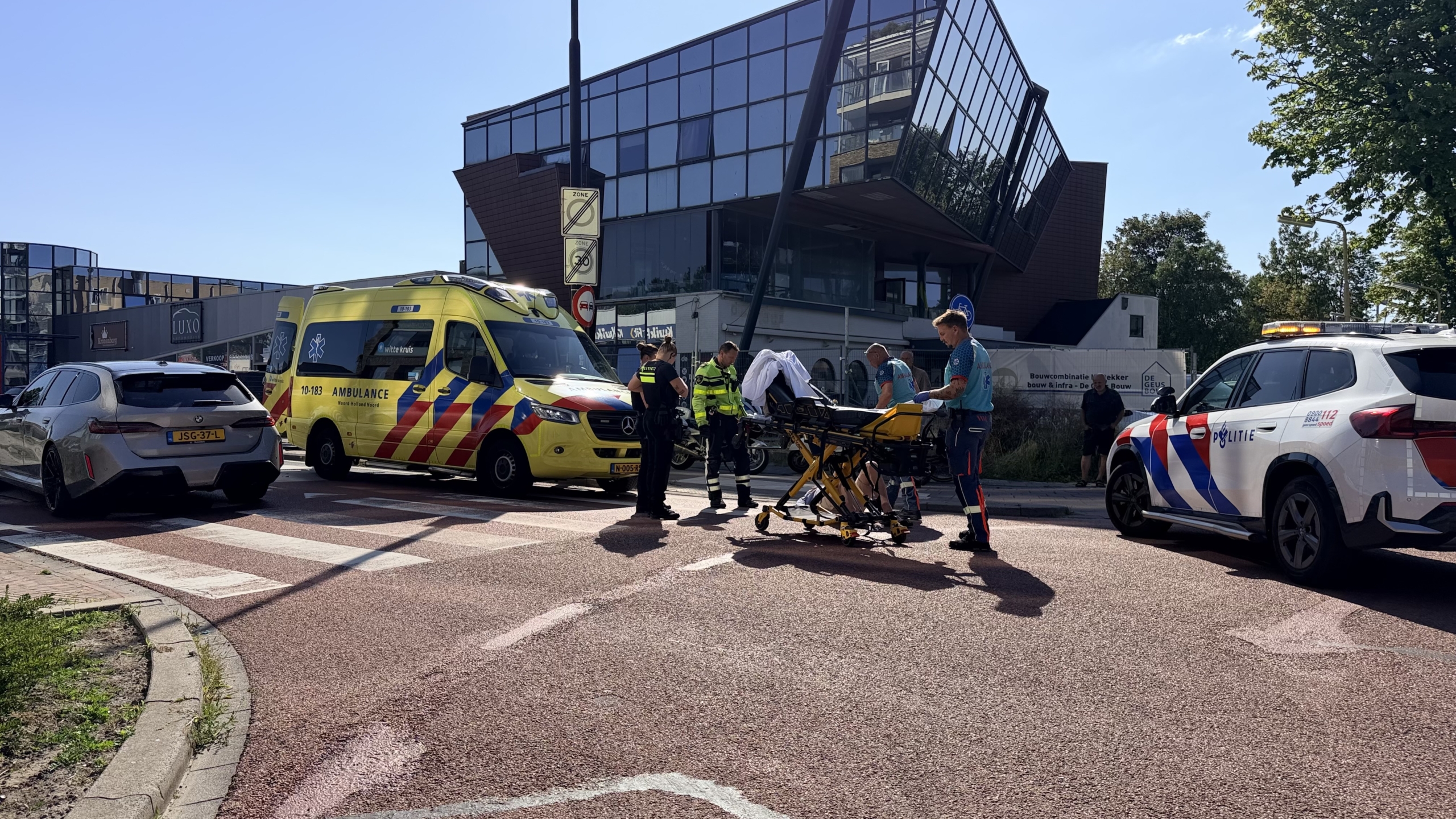 Ambulance en politieauto bij een ongeval, met hulpverleners rond een brancard op een kruispunt voor modern gebouw