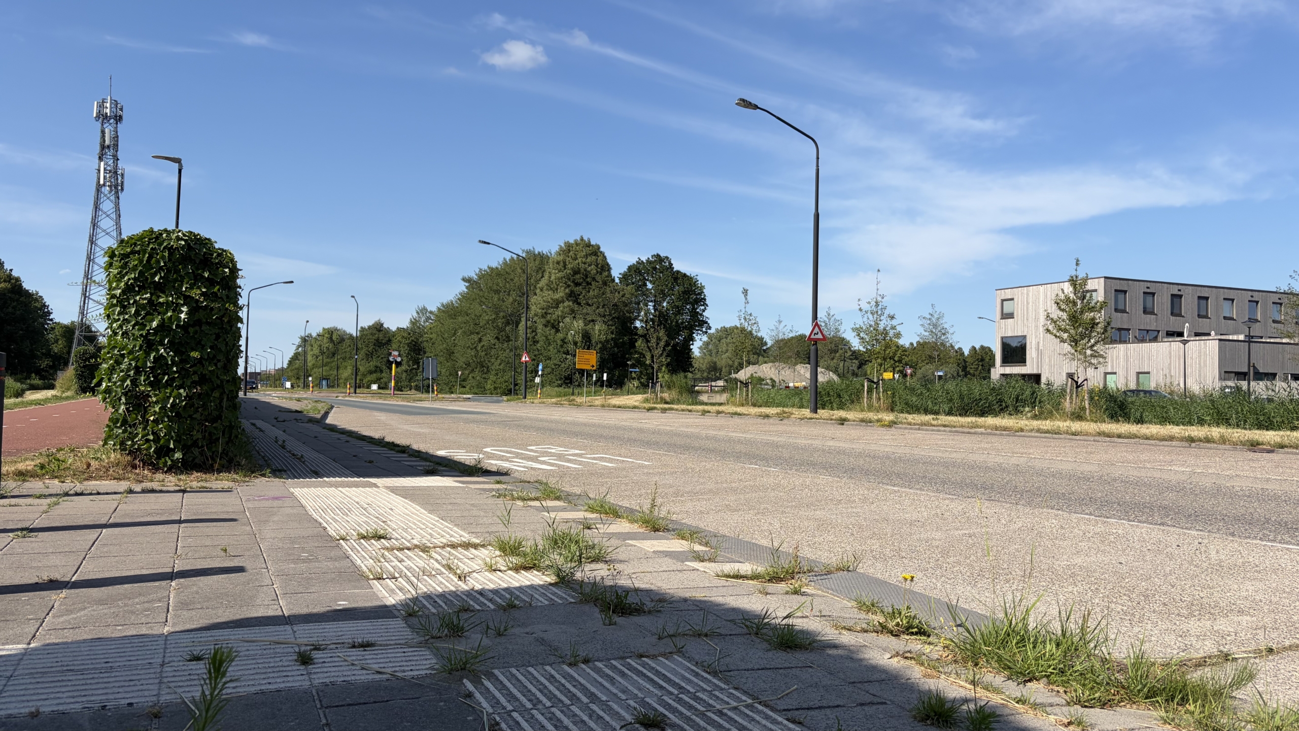 Rustige weg met fietspad, lantaarnpalen en een groen begroeide paal, met kantoorgebouw en zendmast op de achtergrond