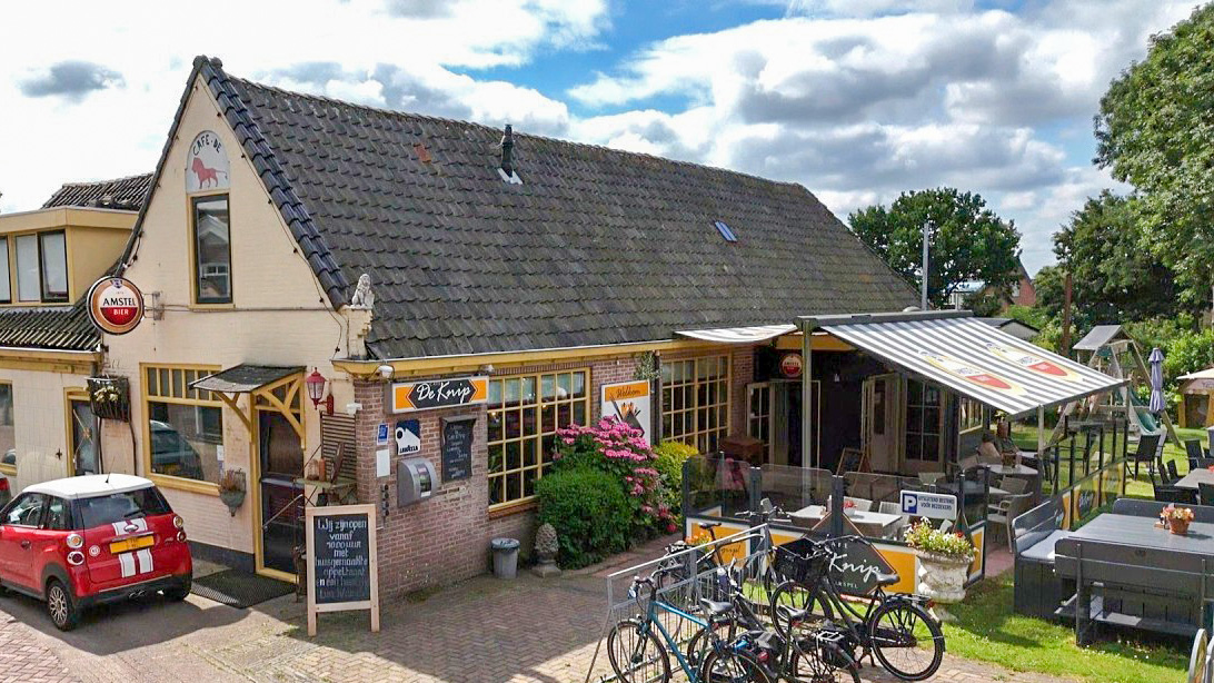 Buitenkant van café De Knijp met terras onder luifel, fietsen voor de deur en een rode auto geparkeerd