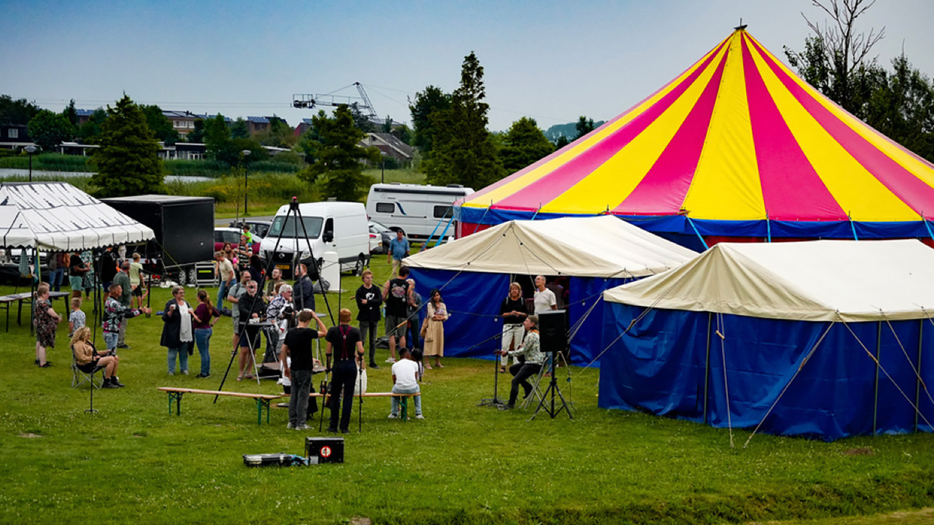 Mensen op een grasveld bij een evenement met een grote geel-roze circustent en blauwe bijtenten