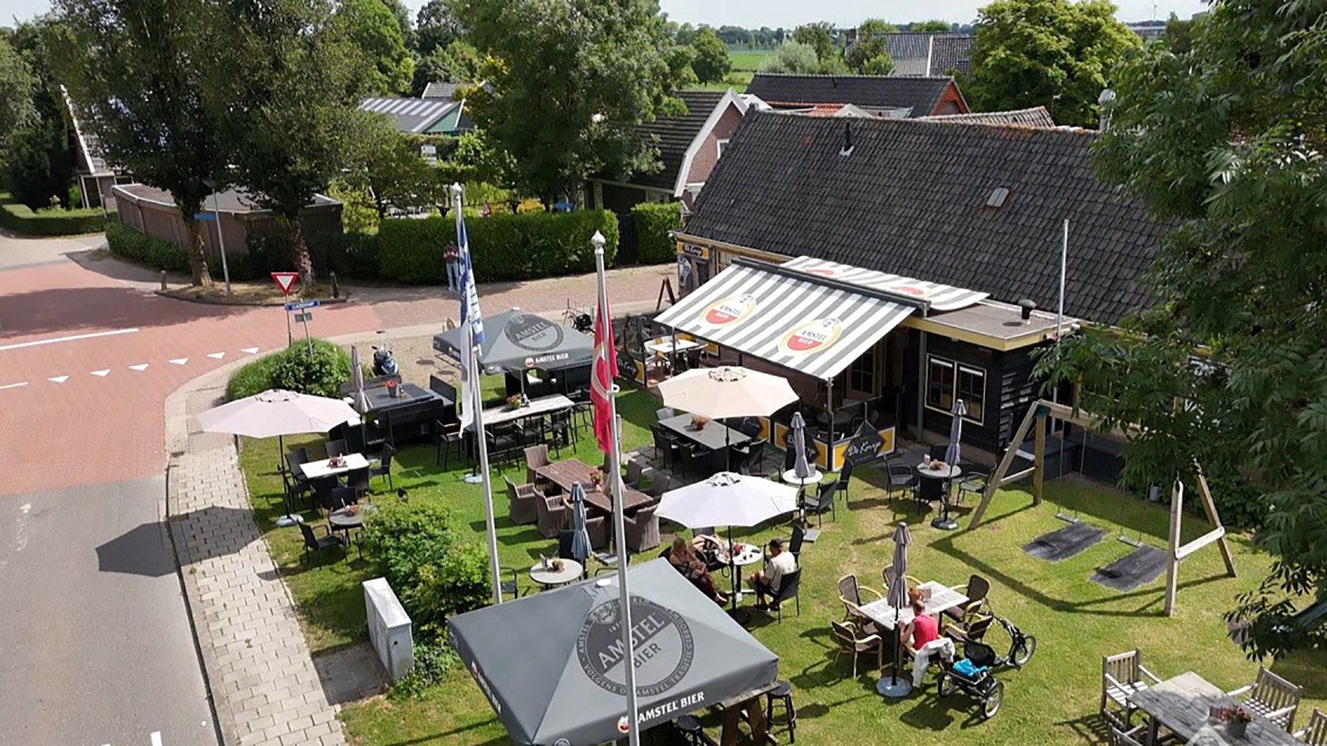 Terras met tafels en parasols naast een café met gestreepte luifel, aan een kruispunt met veel groen