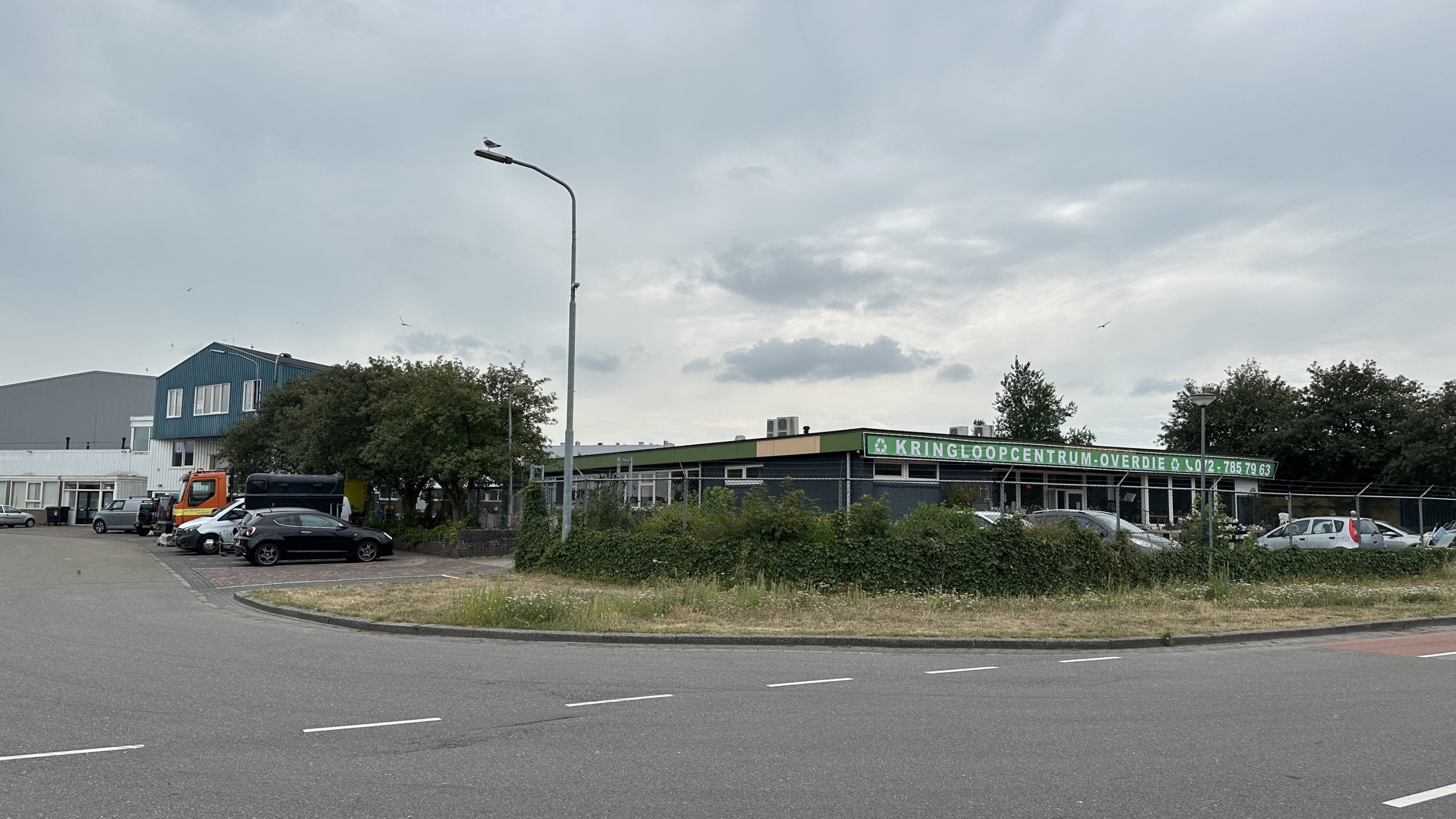 Industrieel gebouw met bord Kringloopcentrum Overdie, met geparkeerde auto’s en een lantaarnpaal langs de weg
