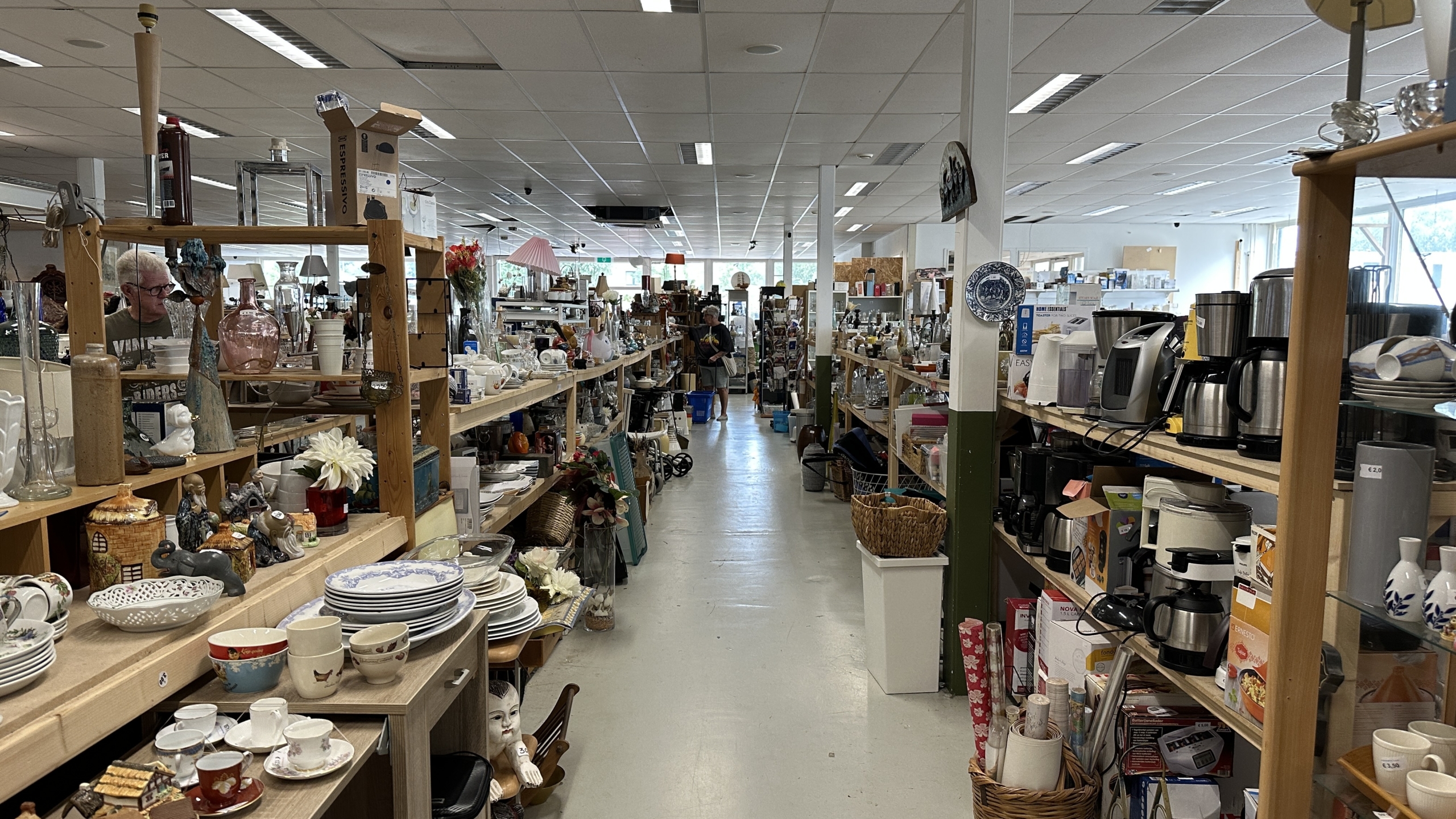 Gangpad in een kringloopwinkel met volle houten stellingen met servies, vazen, lampen en koffiezetapparaten