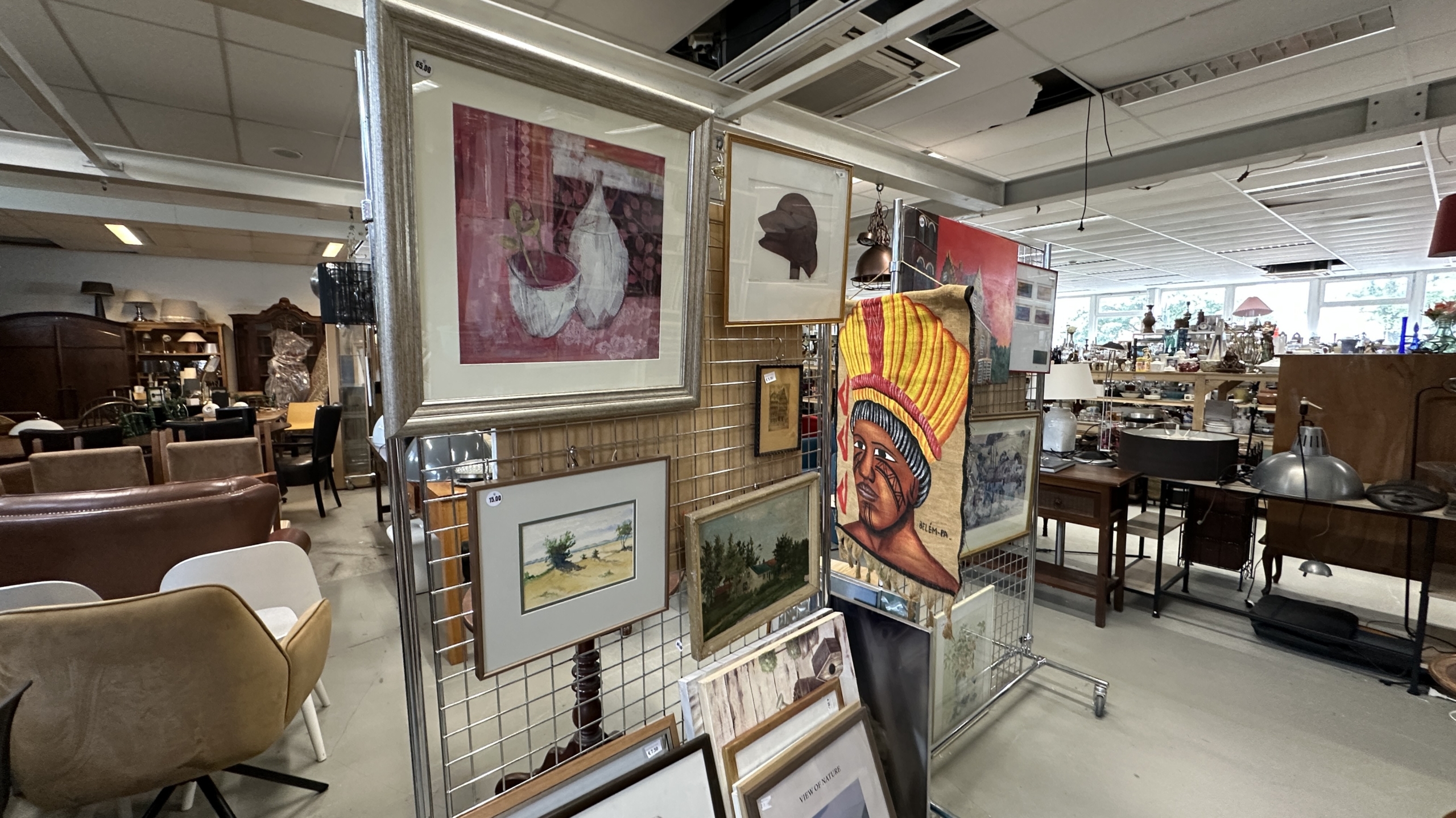 Kunstwerken in lijsten op rekken in een kringloopwinkel, met meubels en lampen op de achtergrond