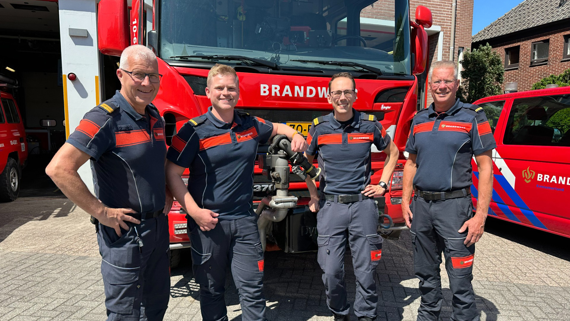 Vier brandweerlieden poseren voor een rode brandweerwagen op de kazerne, met BRAN DWEER zichtbaar op de voorkant