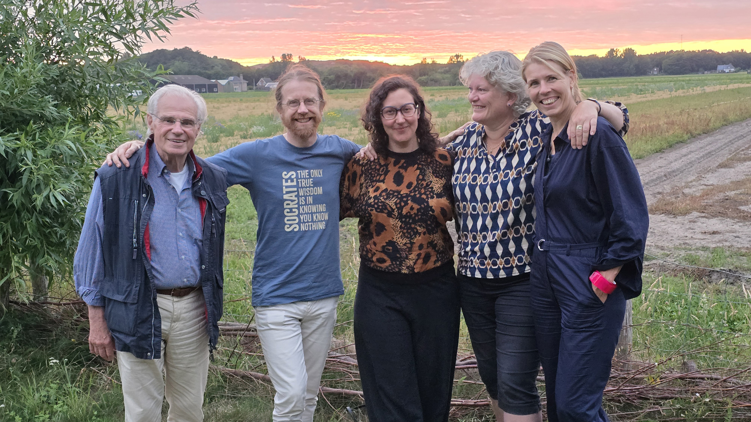 Vijf mensen staan arm in arm op het platteland bij zonsondergang, met velden en een zandpad op de achtergrond
