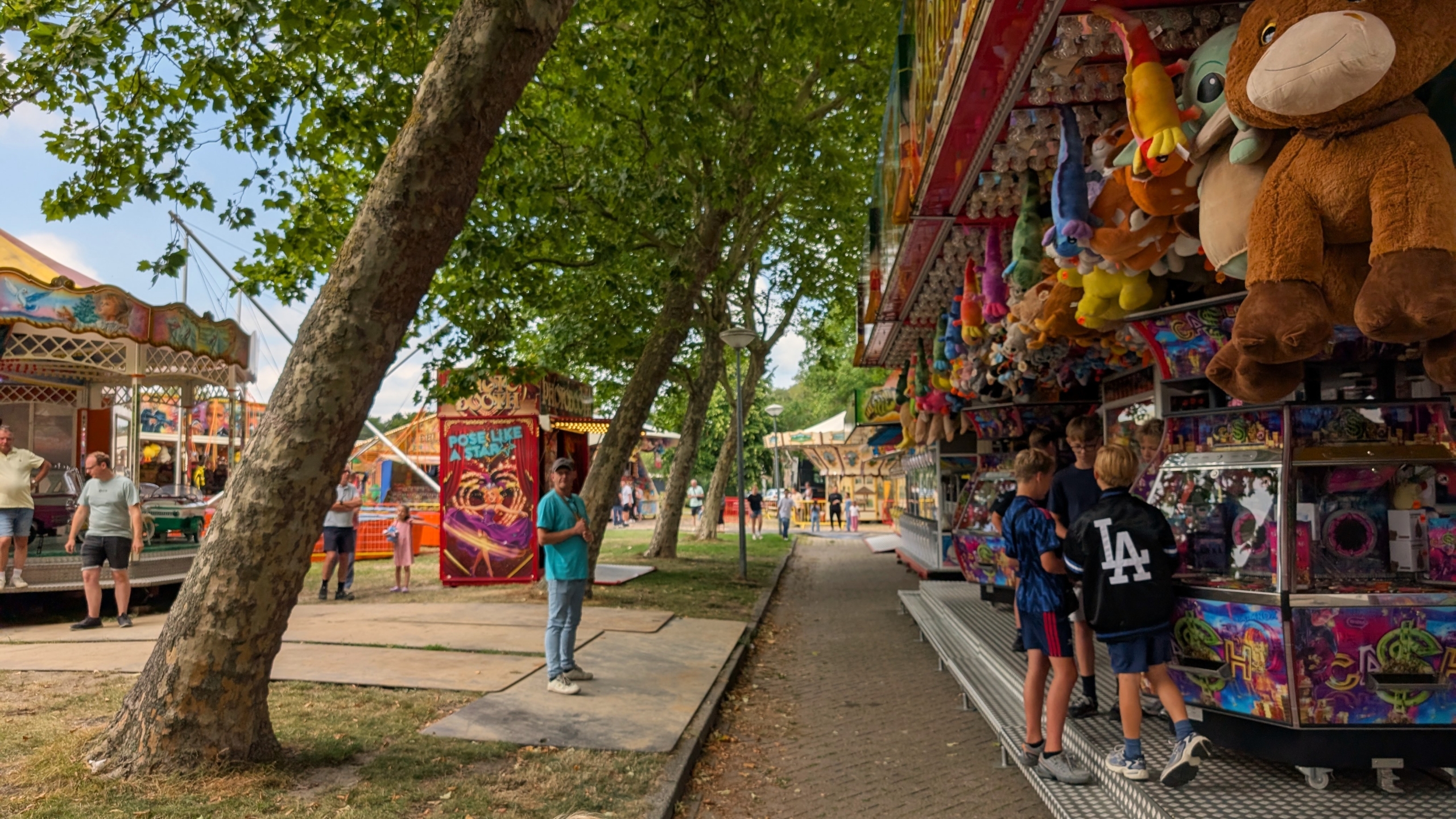 Kermis onder bomen, met bezoekers bij een grijpkraam vol knuffels en attracties op de achtergrond