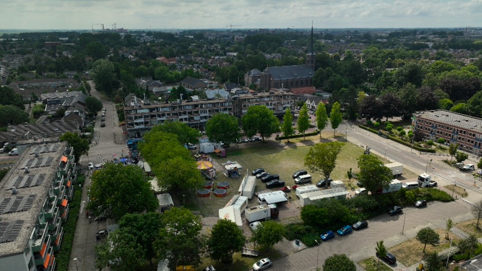 Luchtfoto van woonwijk met open veld met kermisattracties en geparkeerde wagens, met kerk en bomen op de achtergrond