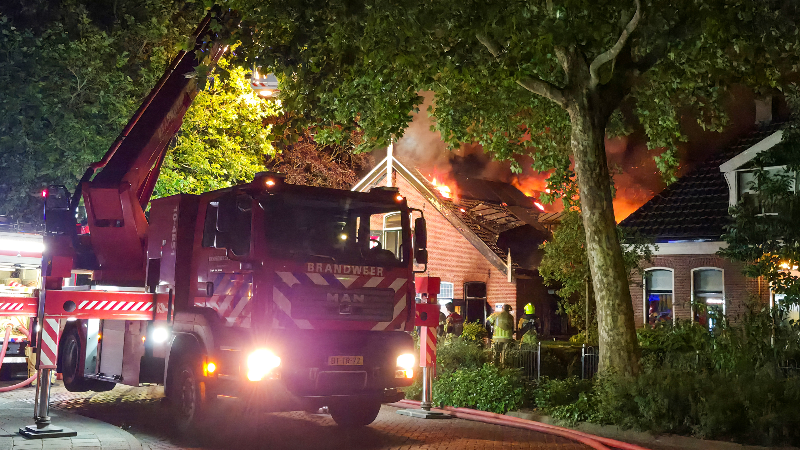 Brandweerwagen met hoogwerker bij uitslaande dakbrand van woning in de nacht, met rook, vlammen en brandweerlieden