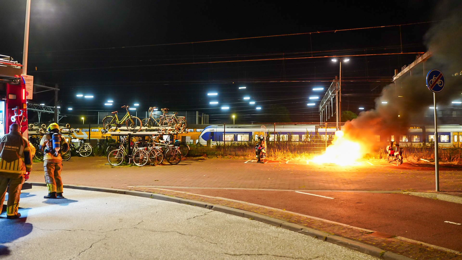 Brand bij fietsenstalling naast een station, met brandweerwagen en brandweerlieden terwijl rook en vlammen opstijgen