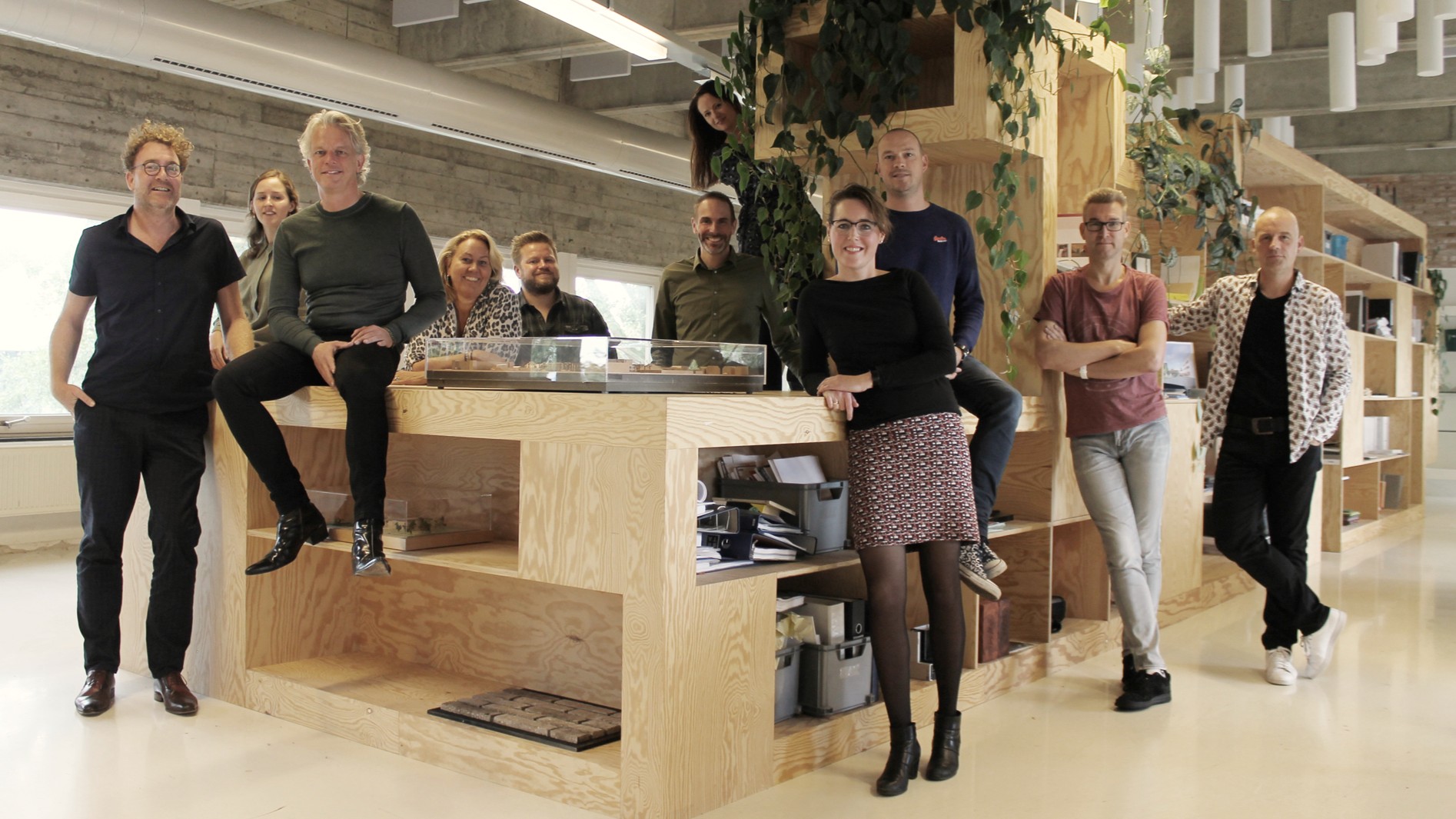 Groepsfoto van tien collega’s in een modern kantoor rond een houten balie met hangplanten en open kasten