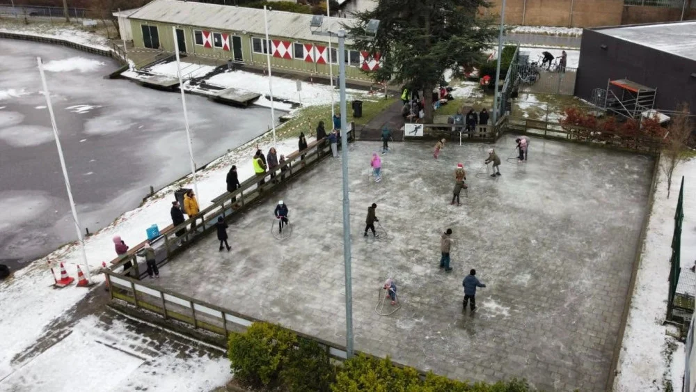 Mensen schaatsen op een omheinde ijsbaan buiten, met sneeuw langs de randen en een deels bevroren water naast de baan