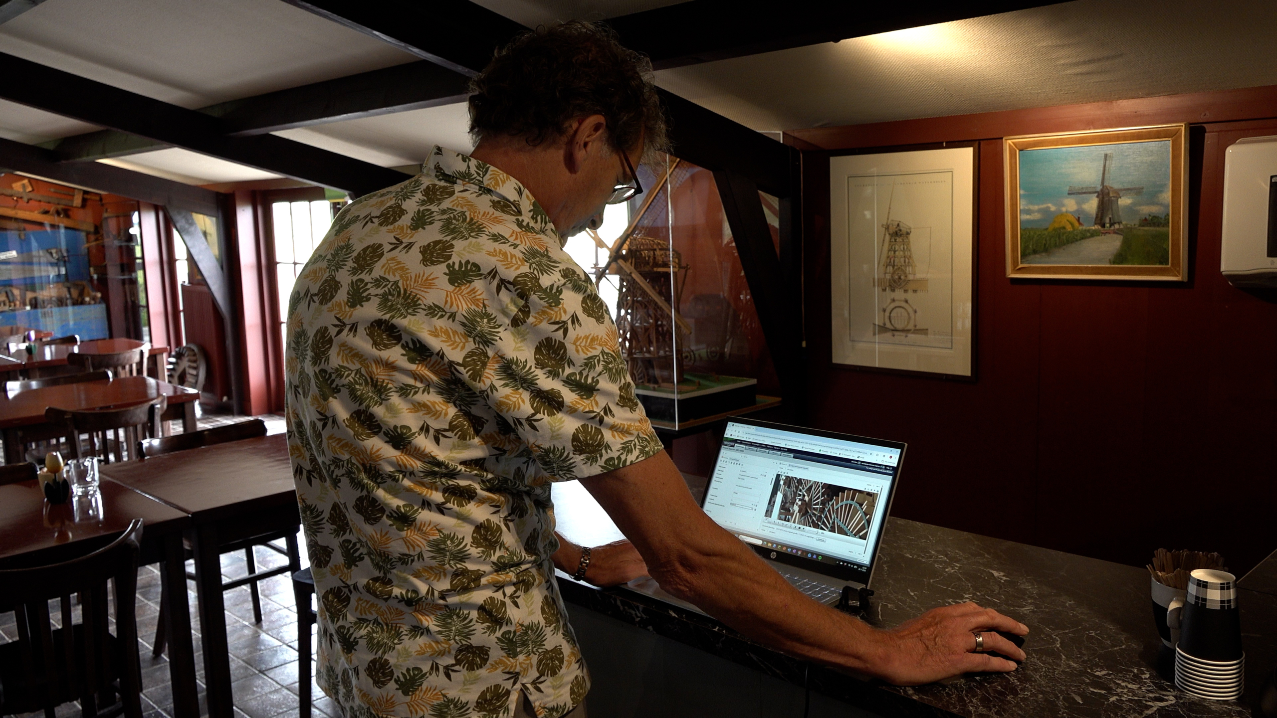 Man in shirt met bladmotief werkt op een laptop aan een balie in een café, met ingelijste molenafbeeldingen aan de muur