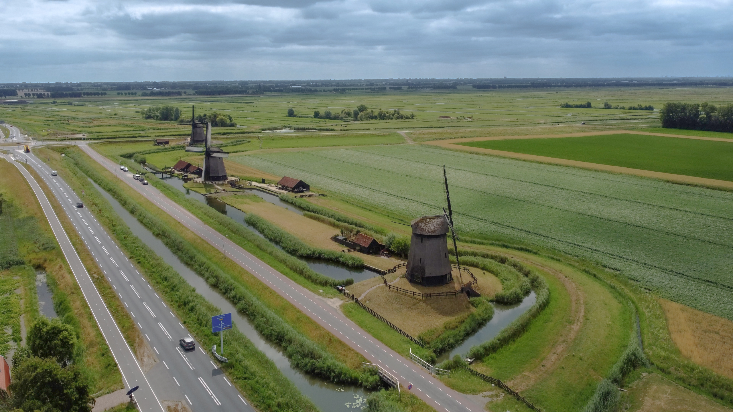 Luchtfoto van twee traditionele windmolens langs een weg en sloot in een uitgestrekt groen polderlandschap
