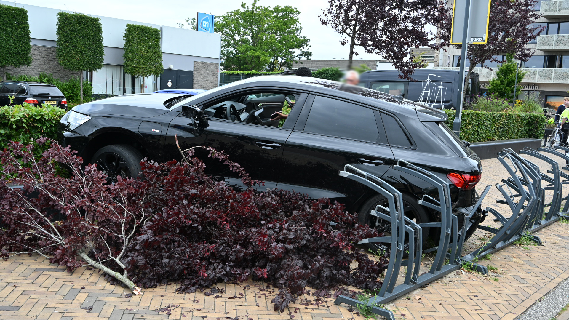 Zwarte auto op stoep tegen fietsenrek gereden, met afgebroken boomtakken ervoor bij een supermarkt