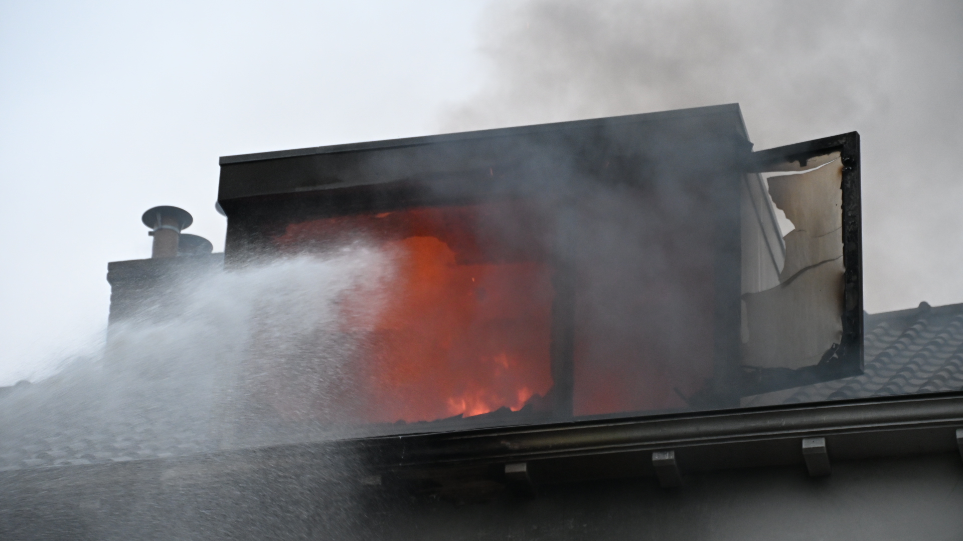 Brand in een dakopbouw met vlammen achter het raam en veel rook, terwijl bluswater tegen het dak spuit