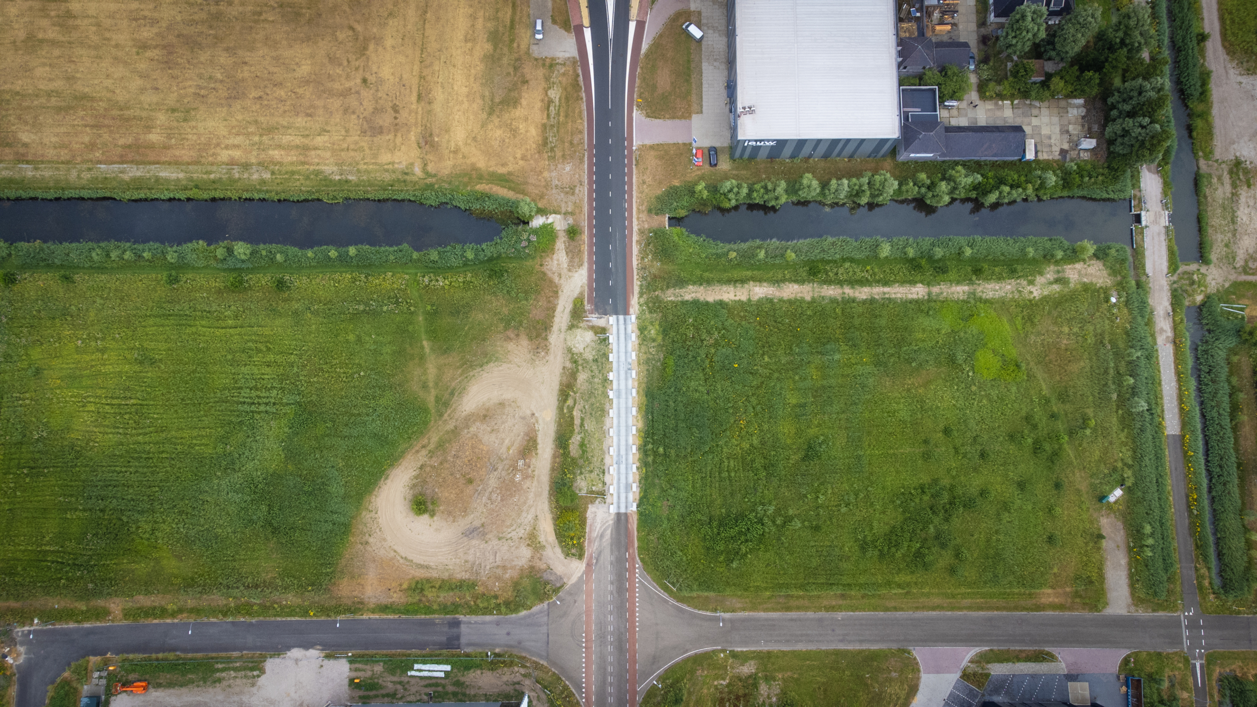 Luchtfoto van een weg met brug over een smalle watergang, omringd door velden en een industriepand